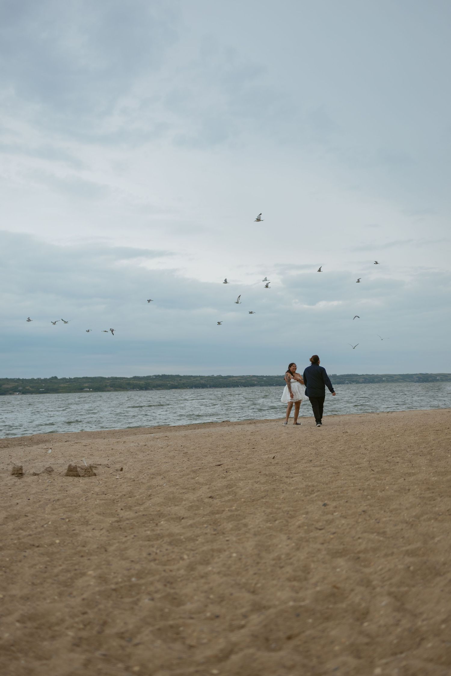 Silhouetted couple walking on beach with seabirds flying overhead against cloudy sky.