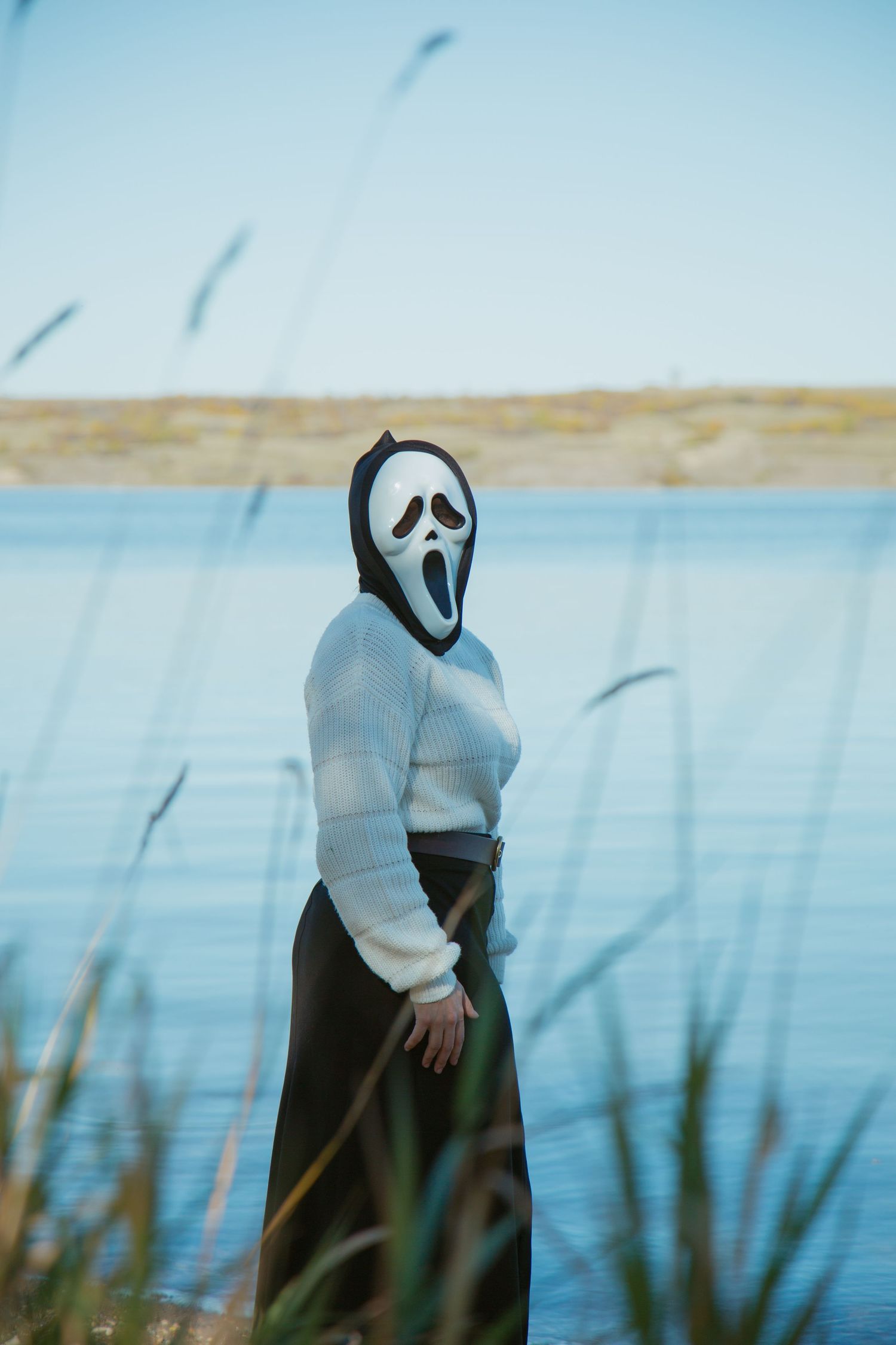 Figure in a white ghost mask stands among tall reeds by a serene lake.