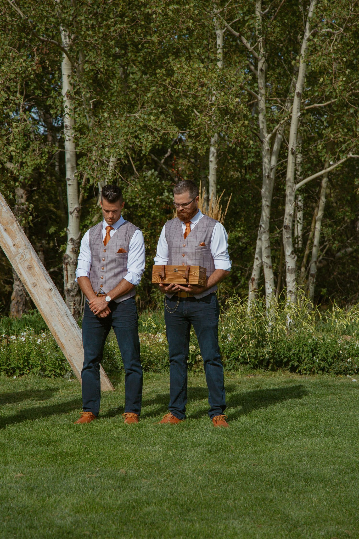 Three people in matching vests and dress pants stand casually on green grass near aspen trees.