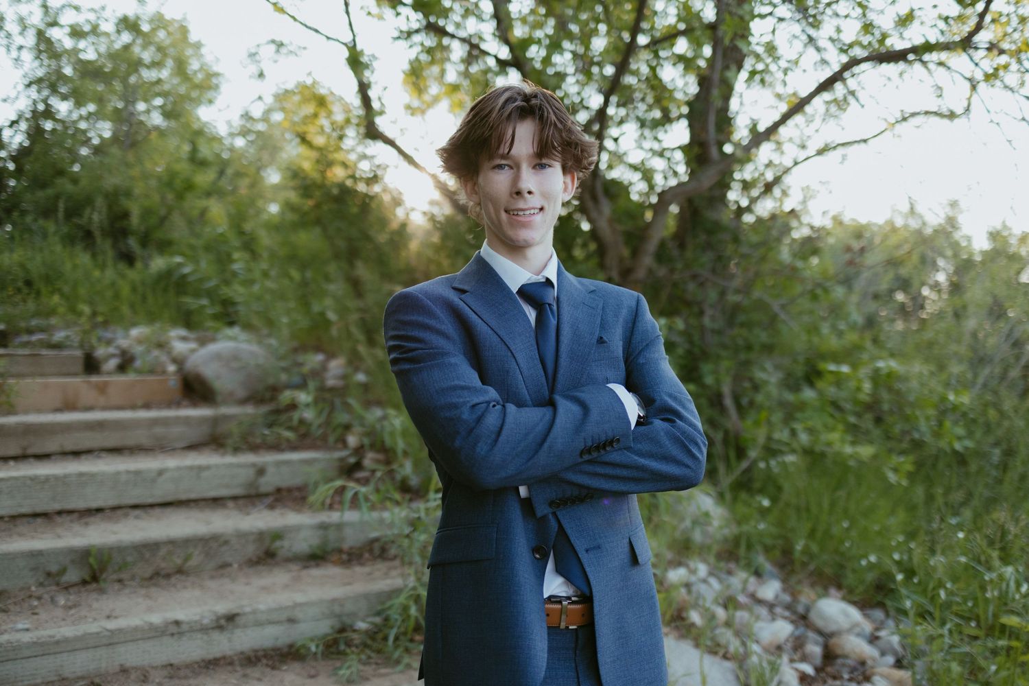 Person in a navy suit sits casually on a wooden bench surrounded by lush green foliage.