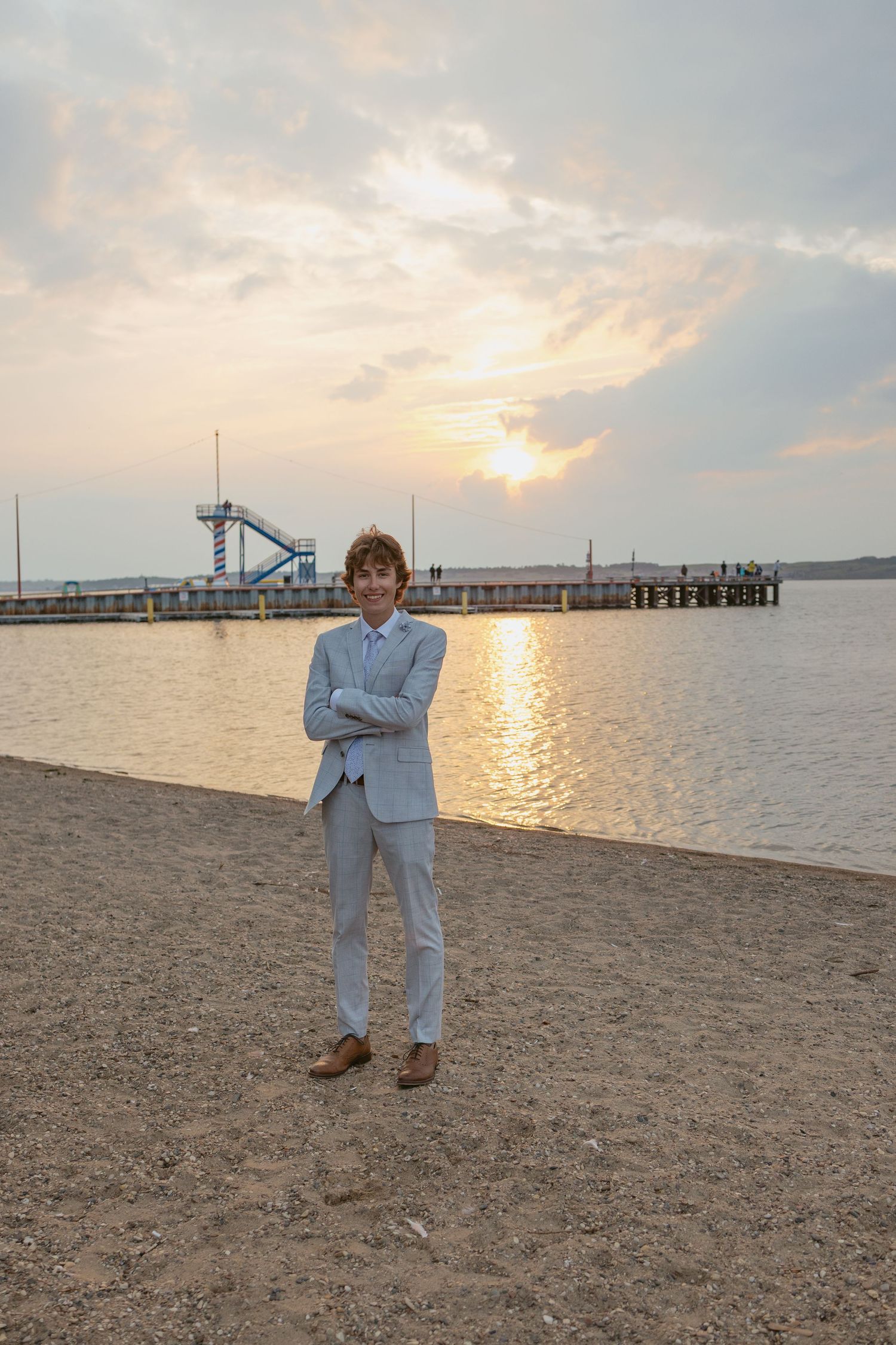Person in light suit poses on beach at sunset with pier and water slide in background.