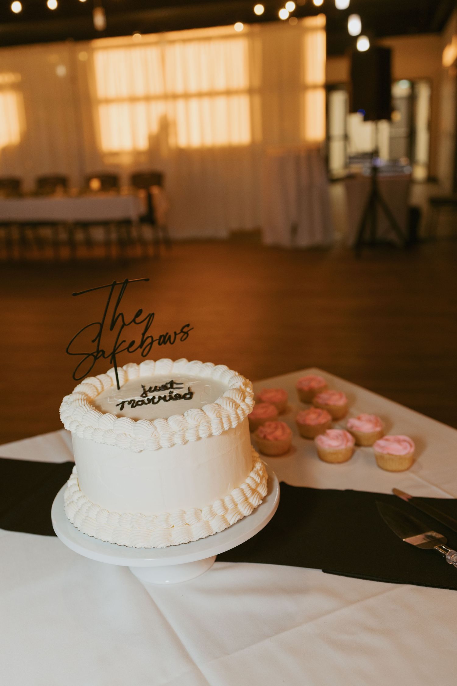 White wedding cake with script topper on cake stand beside pink cupcakes at elegant reception venue.