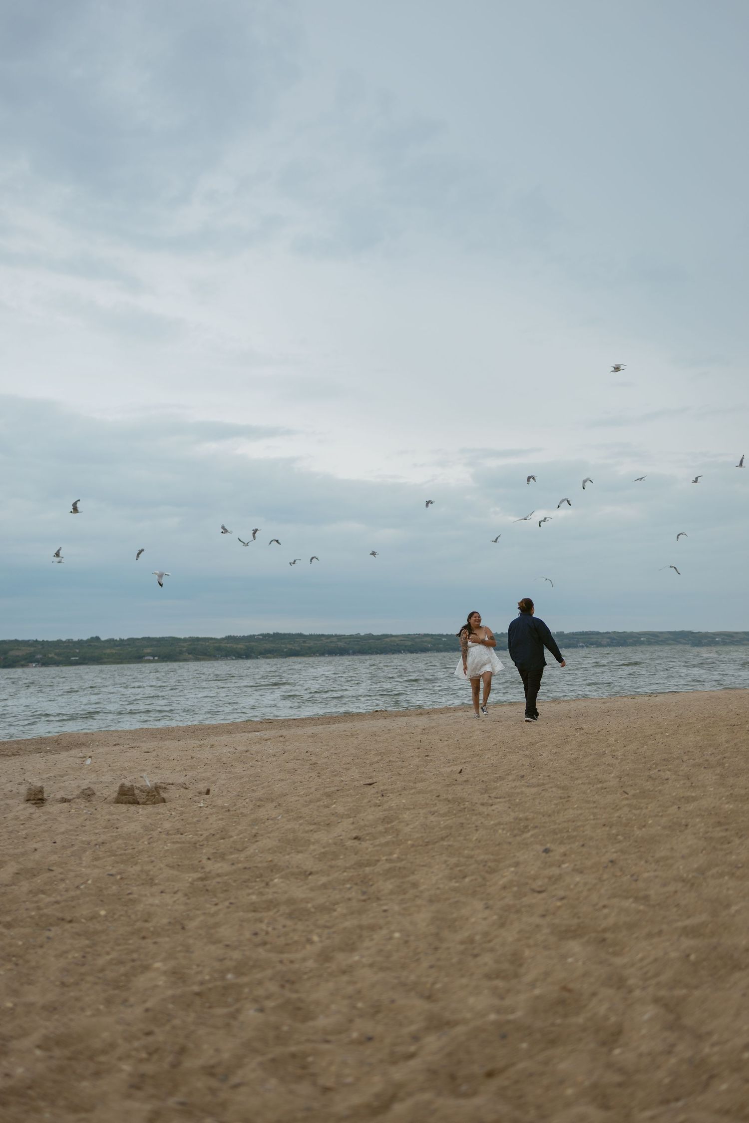 Couple walking on beach holding hands while seagulls fly overhead against cloudy sky.