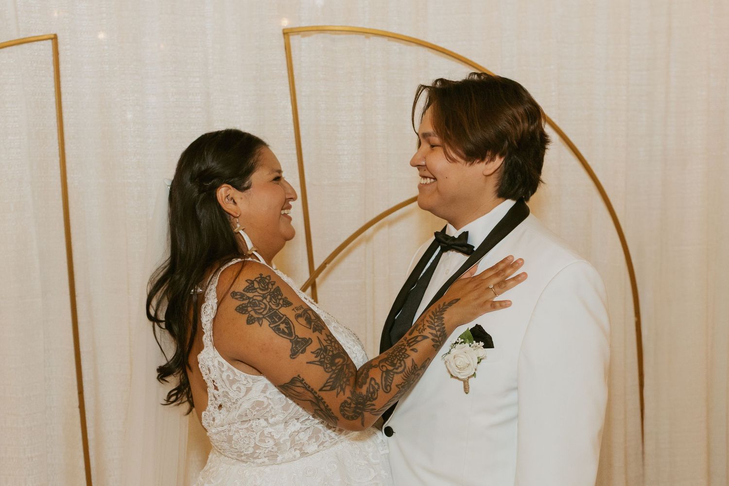 Bride and groom in white wedding attire smile at each other in front of a gold geometric backdrop.