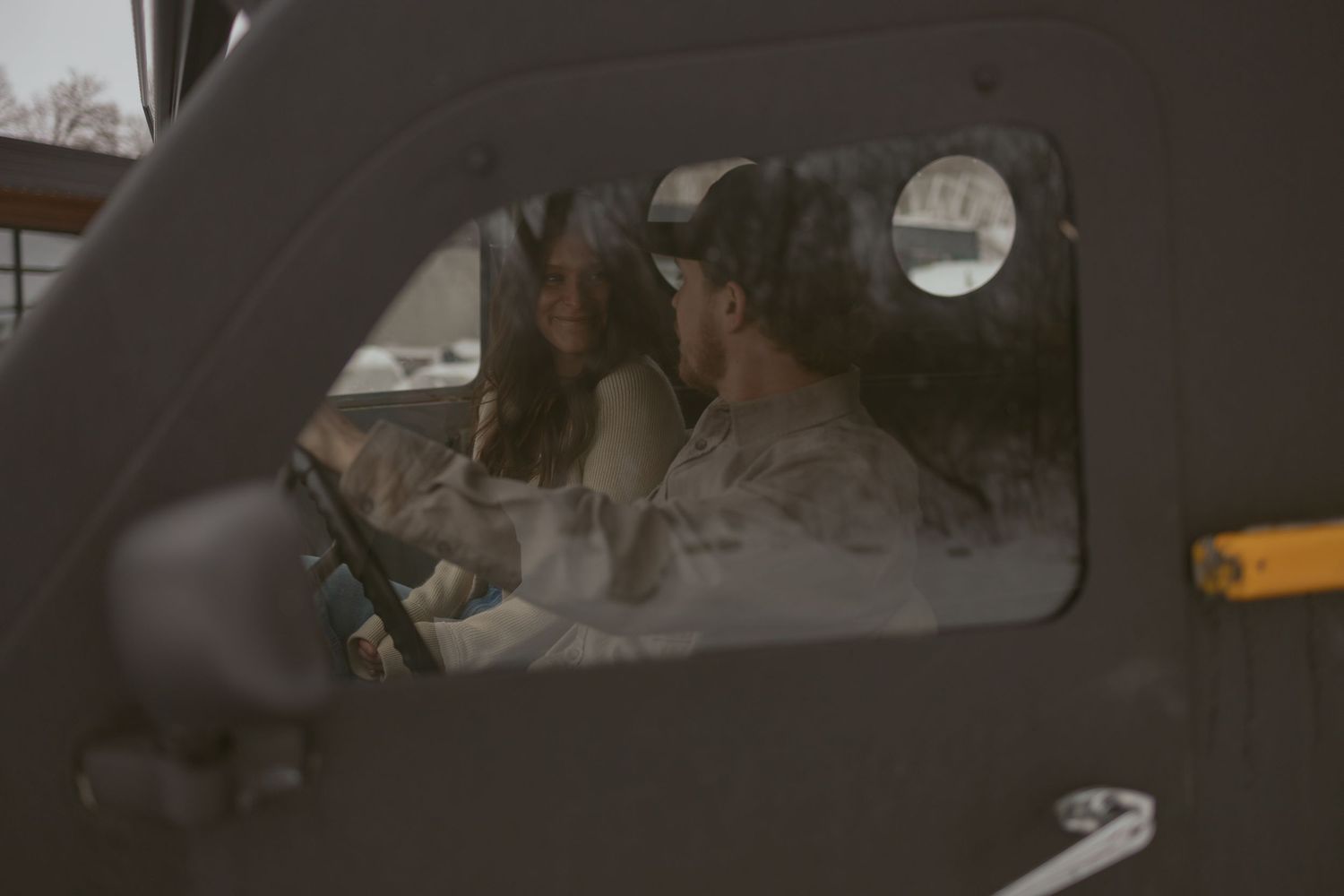 Couple sitting together inside vintage truck cab viewed through side window.