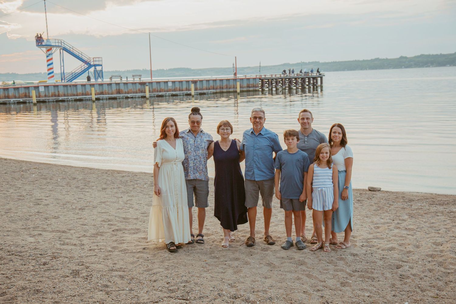 Family group poses on beach at sunset with pier and water slide visible in background.