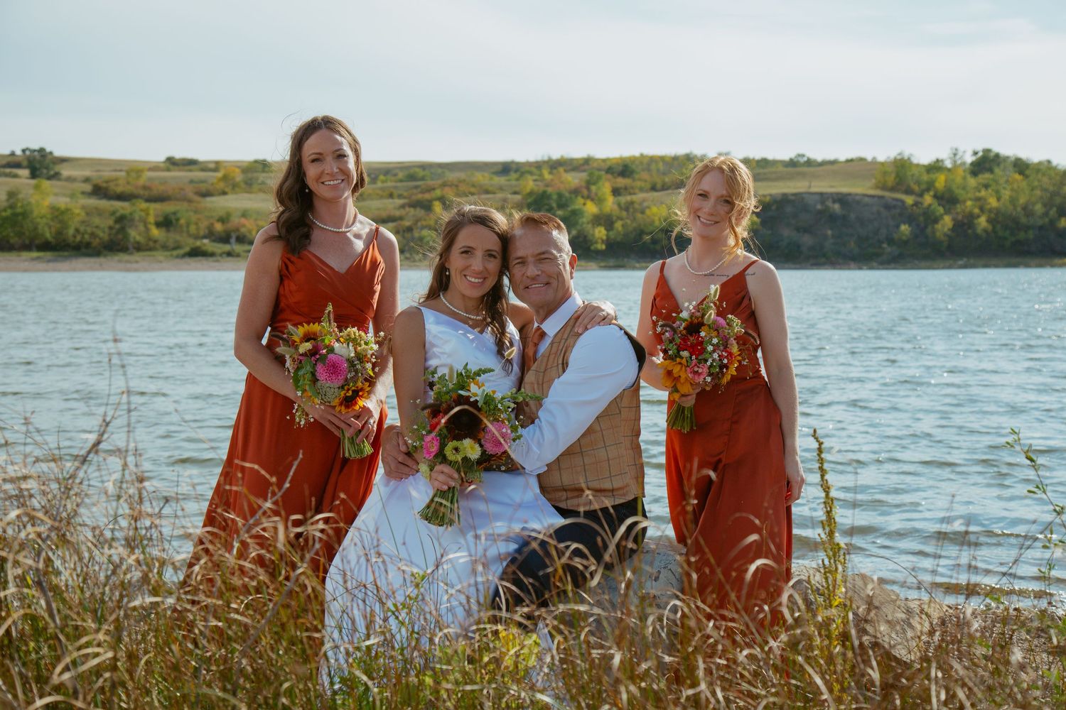 Wedding party in white and rust-colored dresses poses by a lakeside on a sunny day.