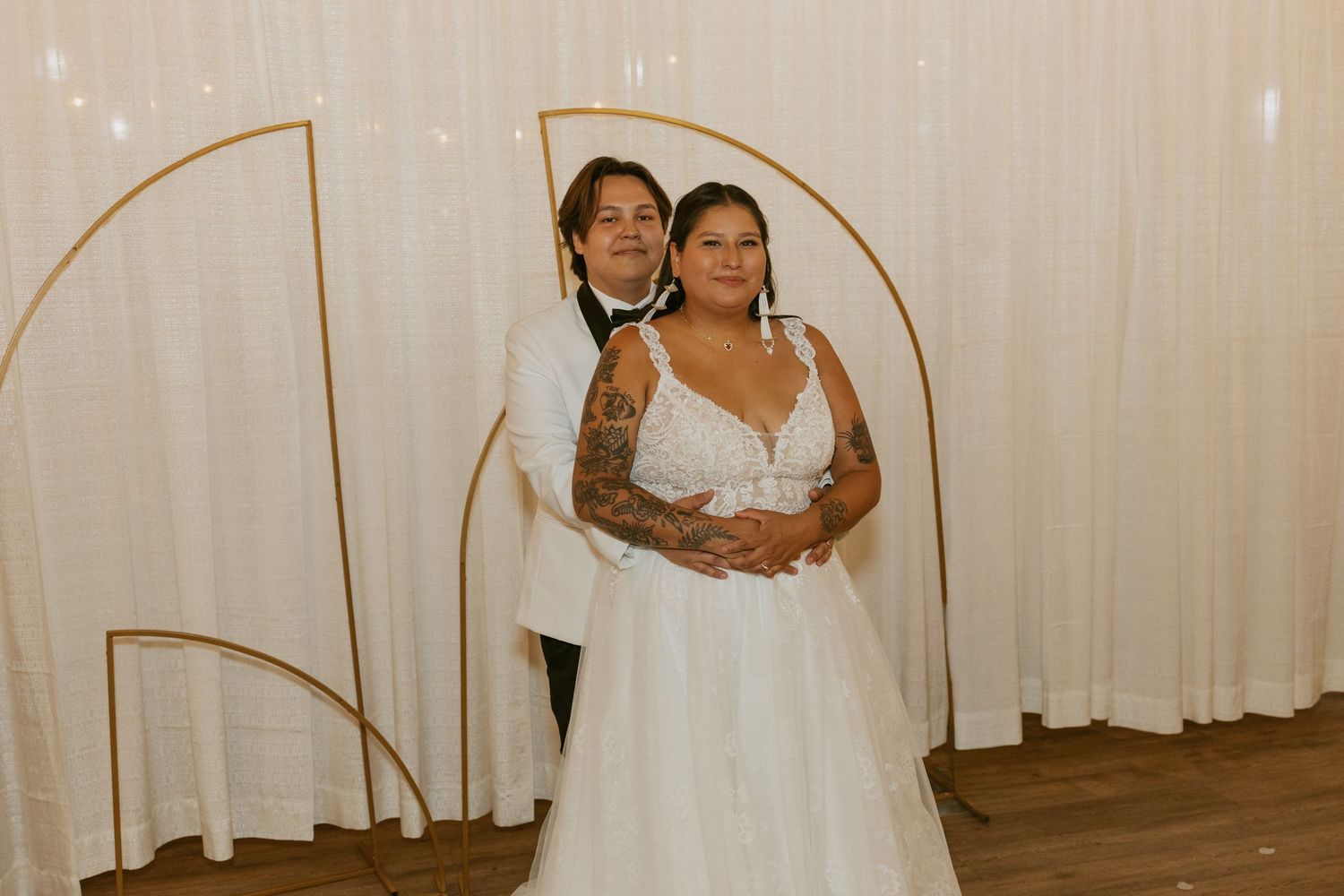 Two brides pose together in wedding attire, one in a white gown and one in a white suit, before a gold arch backdrop.