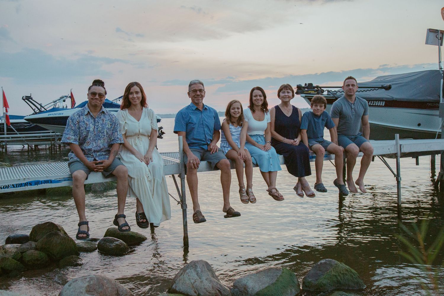 Family group sits casually on rocky shoreline at sunset with boats in background.