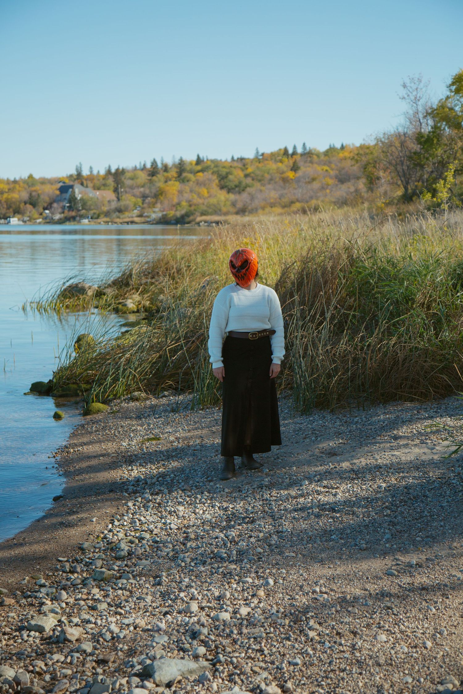 Person in white sweater and long skirt standing by a lakeside path during autumn.