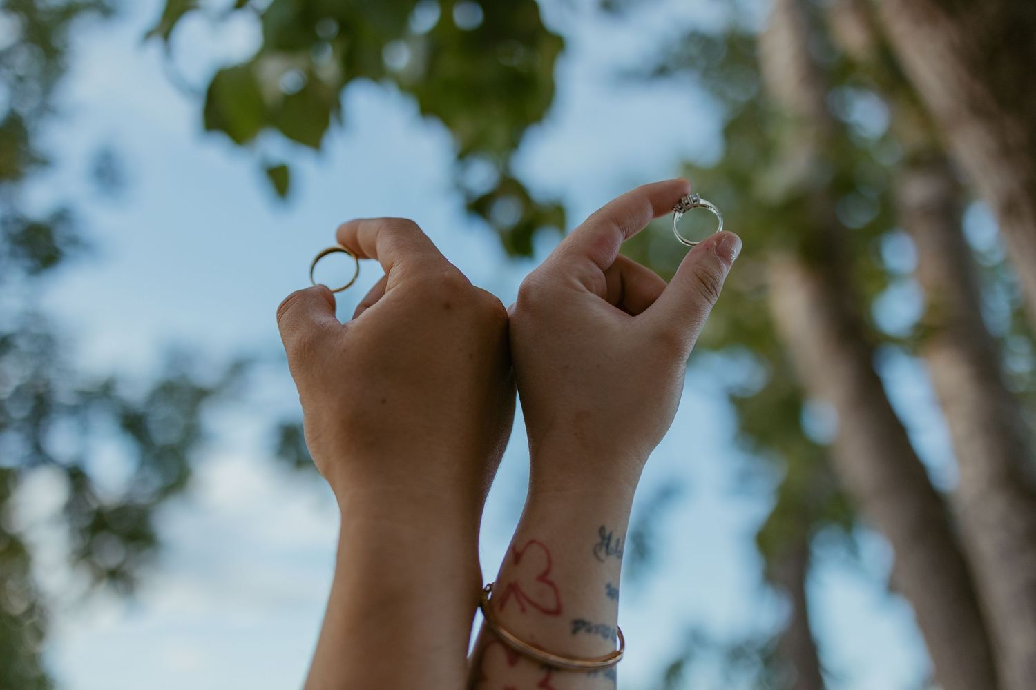 Hands raised against blue sky with trees showing engagement rings.