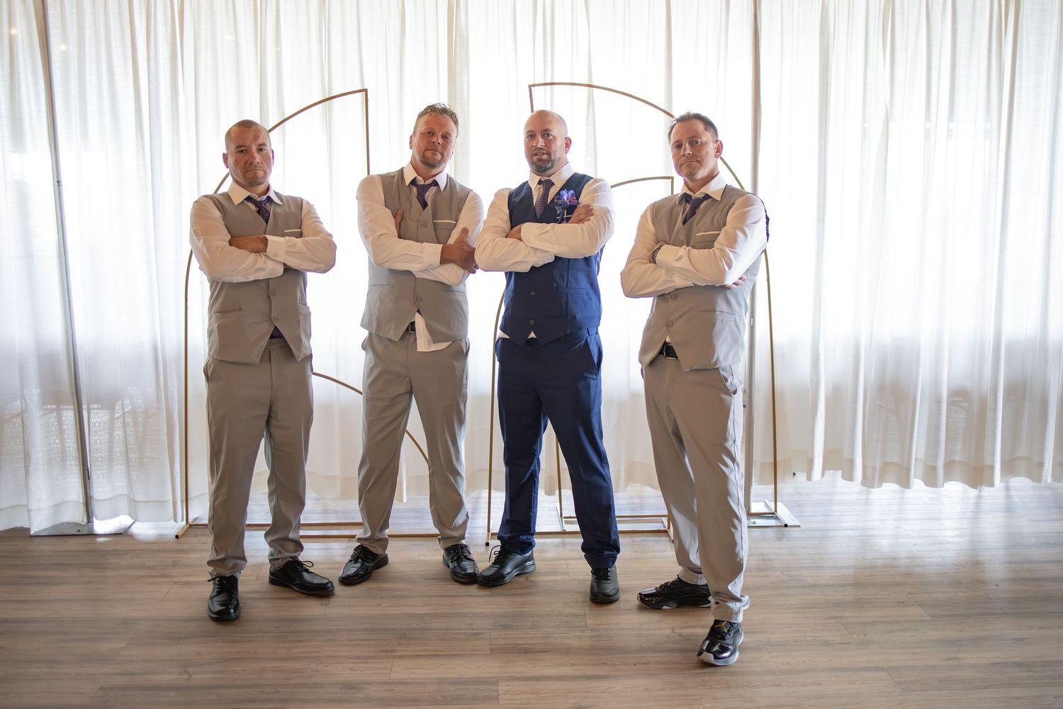 Groom in blue suit stands with three groomsmen in tan suits, arms crossed, posing in front of white curtains.