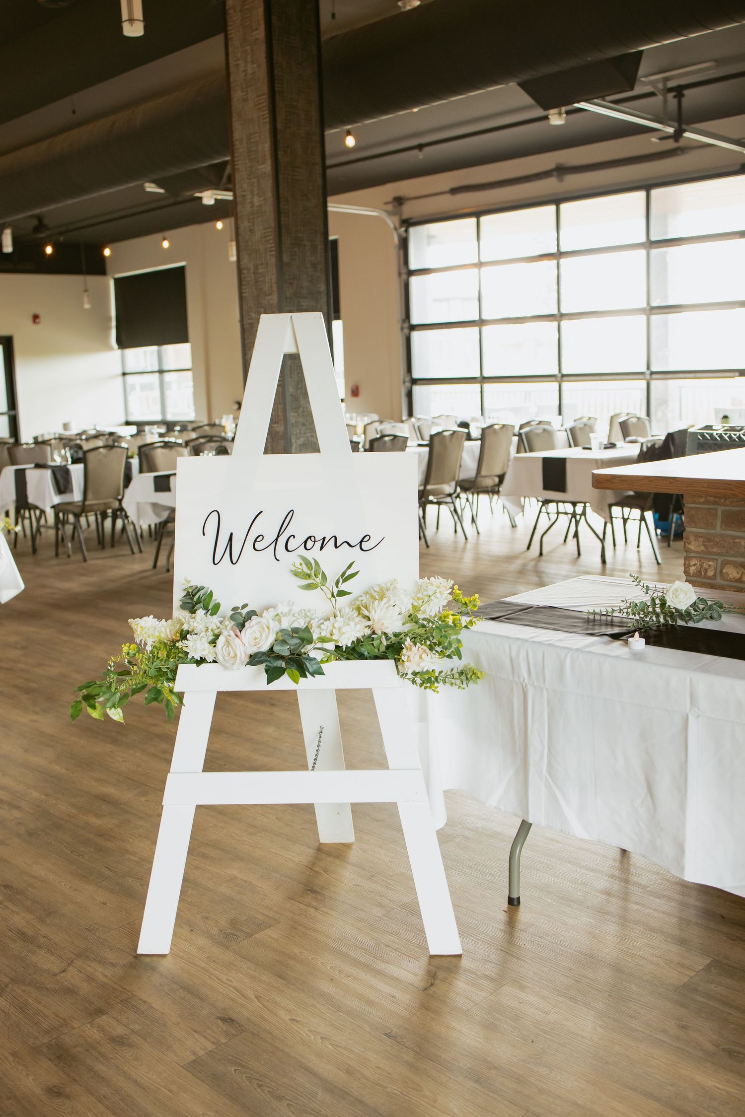 White easel welcome sign with floral decor at a wedding reception venue with tables and chairs.