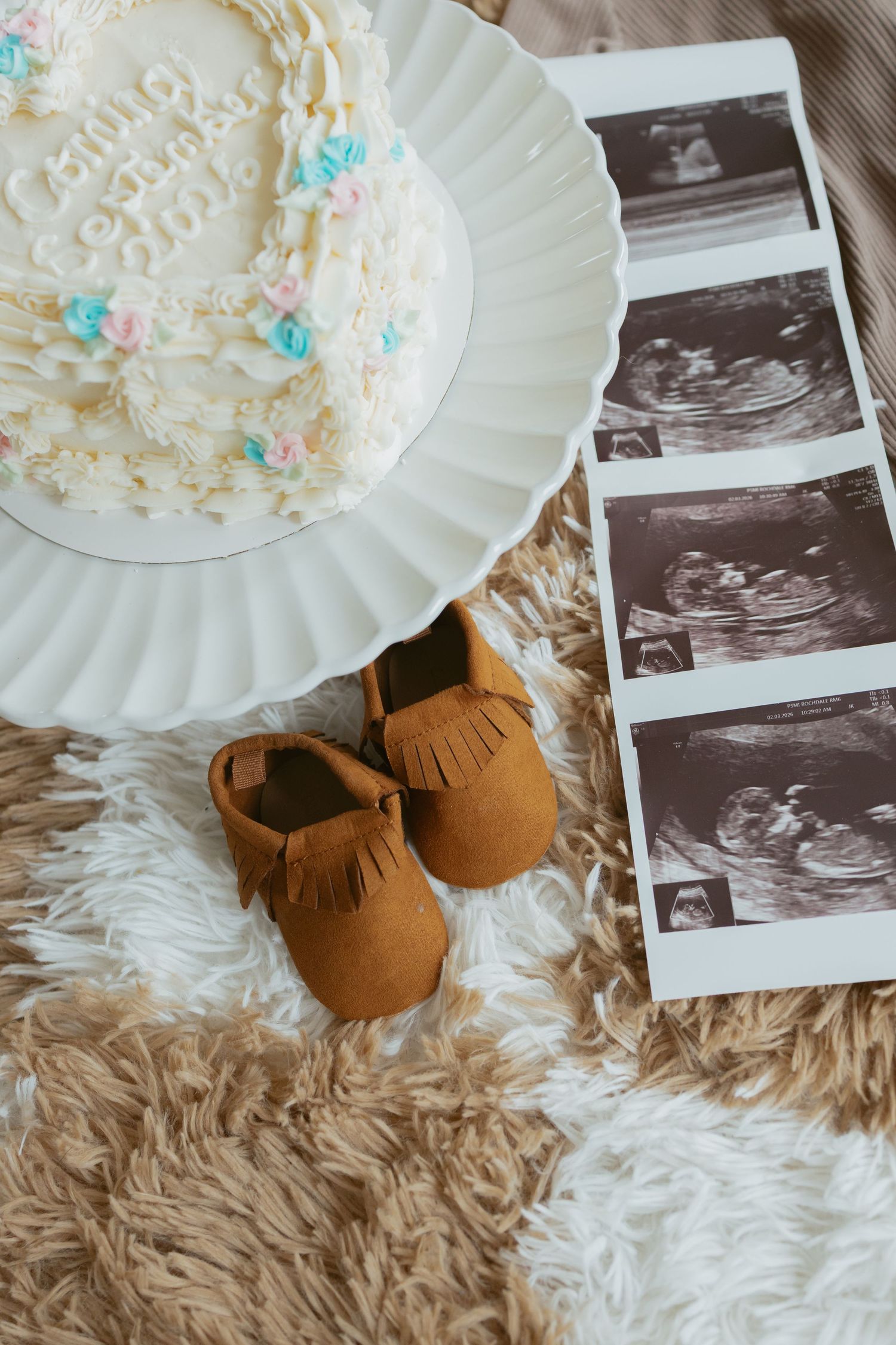 Baby shower flat lay with ultrasound photos, tan suede moccasins, and white gender reveal cake on fluffy rug.