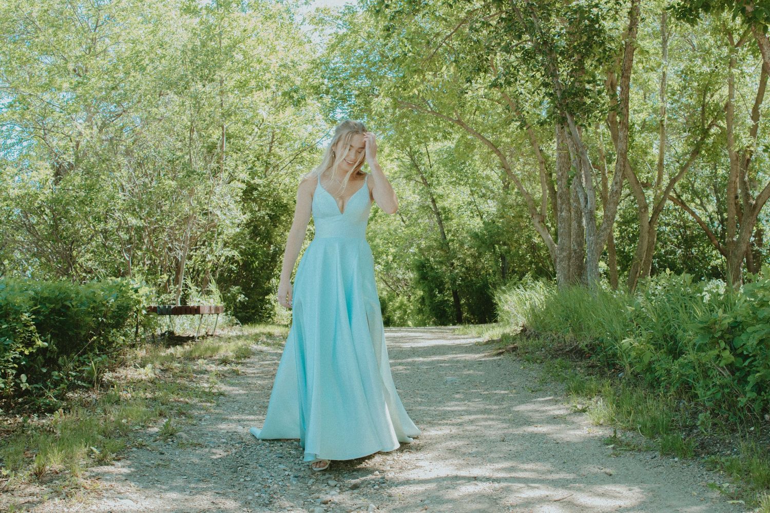 A woman in a flowing mint green gown walks along a sunlit forest path surrounded by lush green trees.