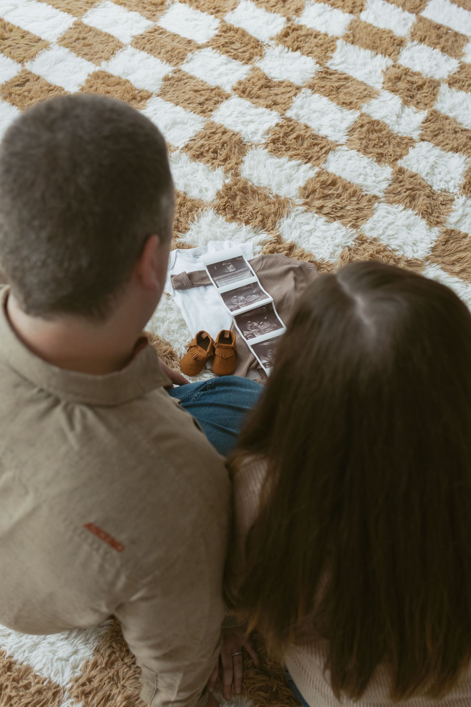 Couple sitting on checkered rug viewing ultrasound photos and tiny baby shoes, seen from above.