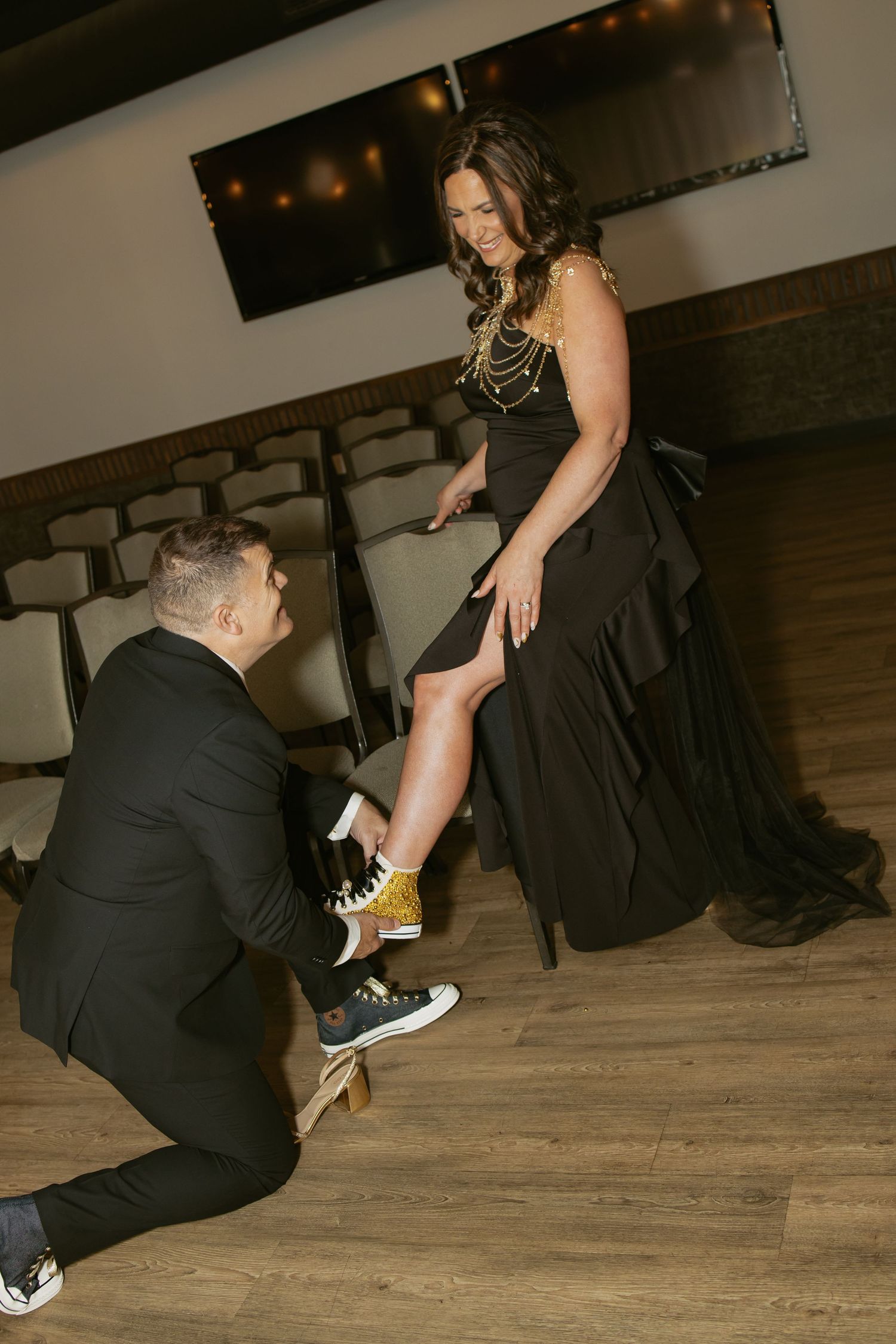 Man kneeling to help woman in black gown with gold sneakers in elegant venue.