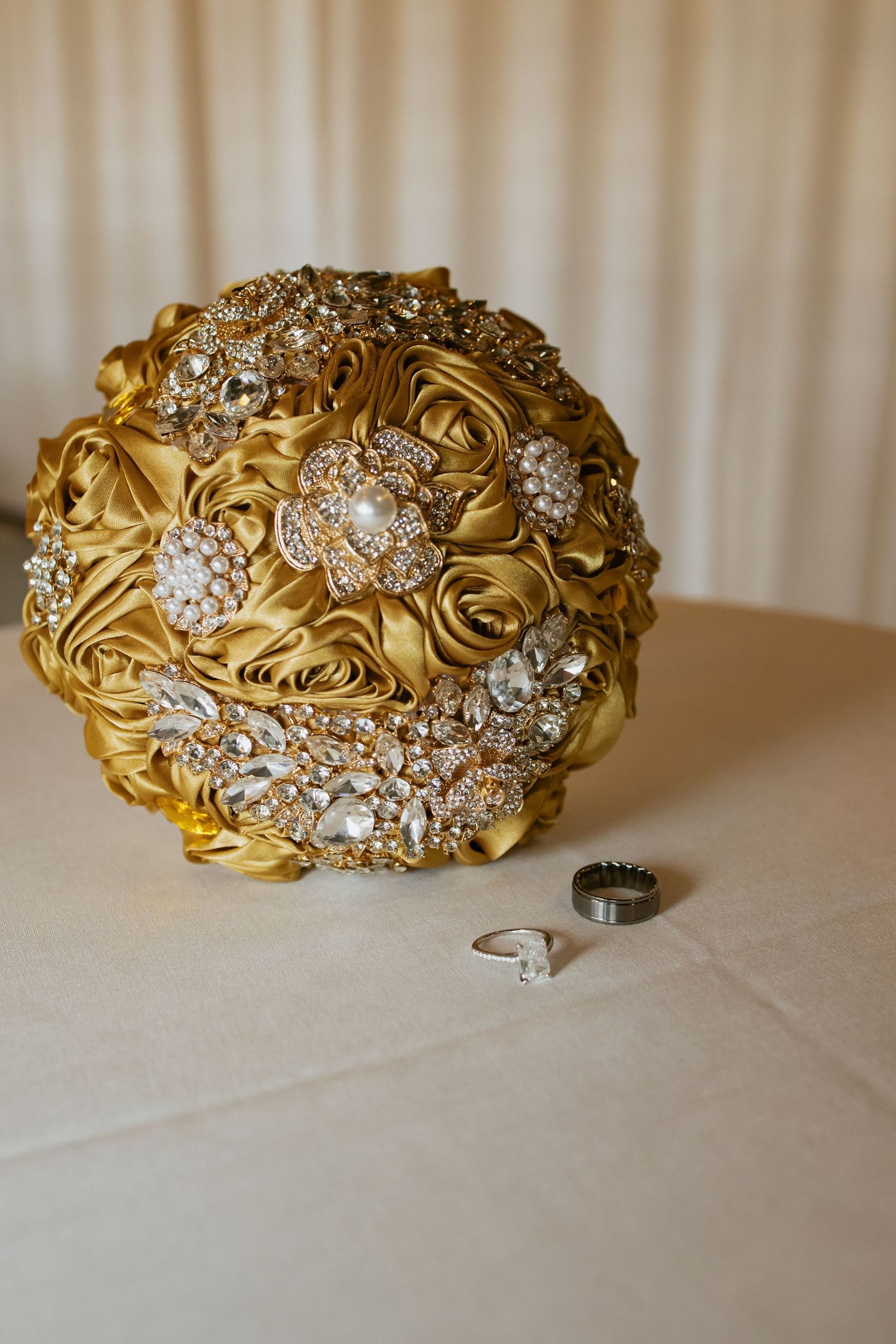 Gold satin brooch bouquet adorned with crystal jewels beside two wedding rings on a white tablecloth.