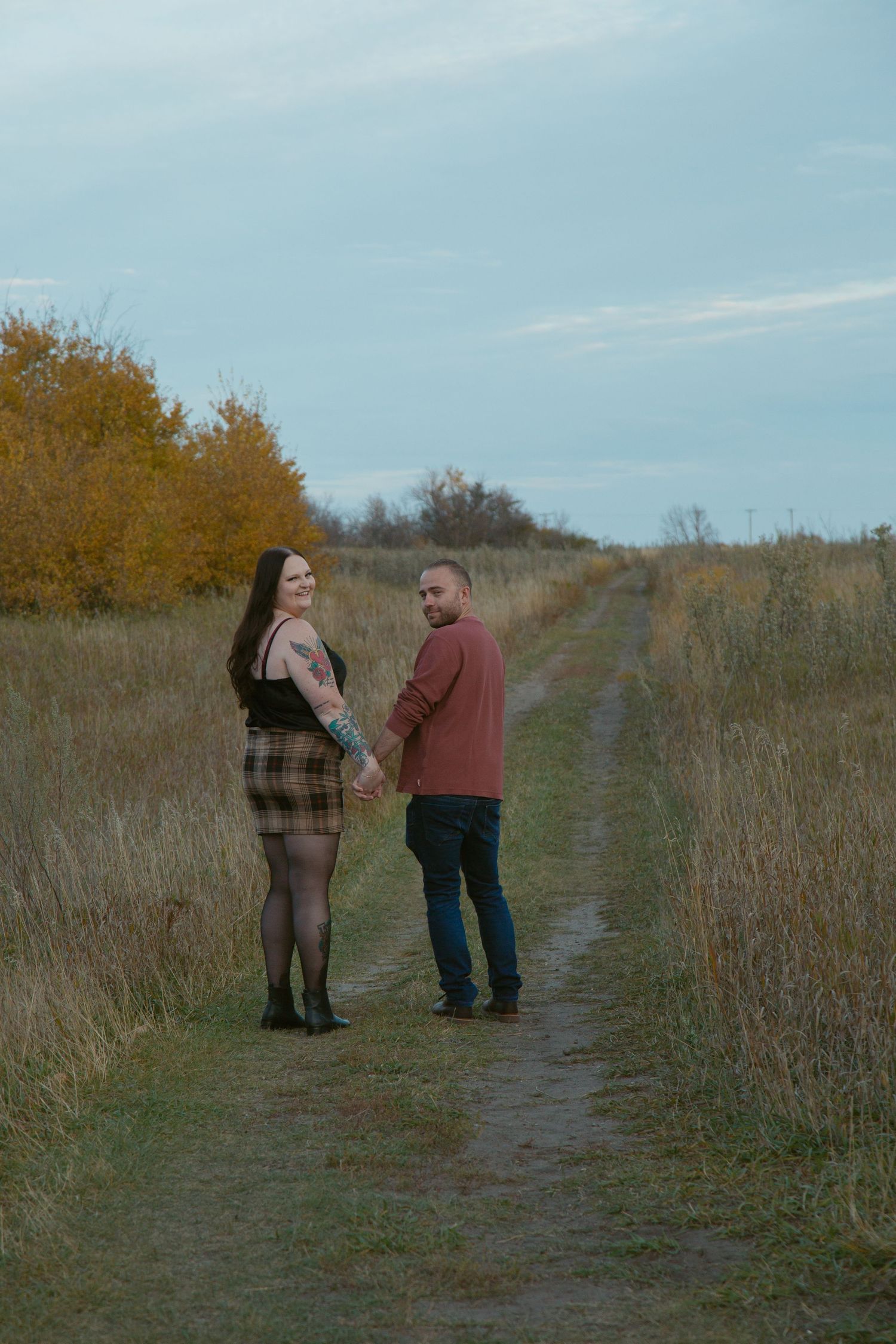 A couple stands together on a path through autumn prairie grass under blue skies.