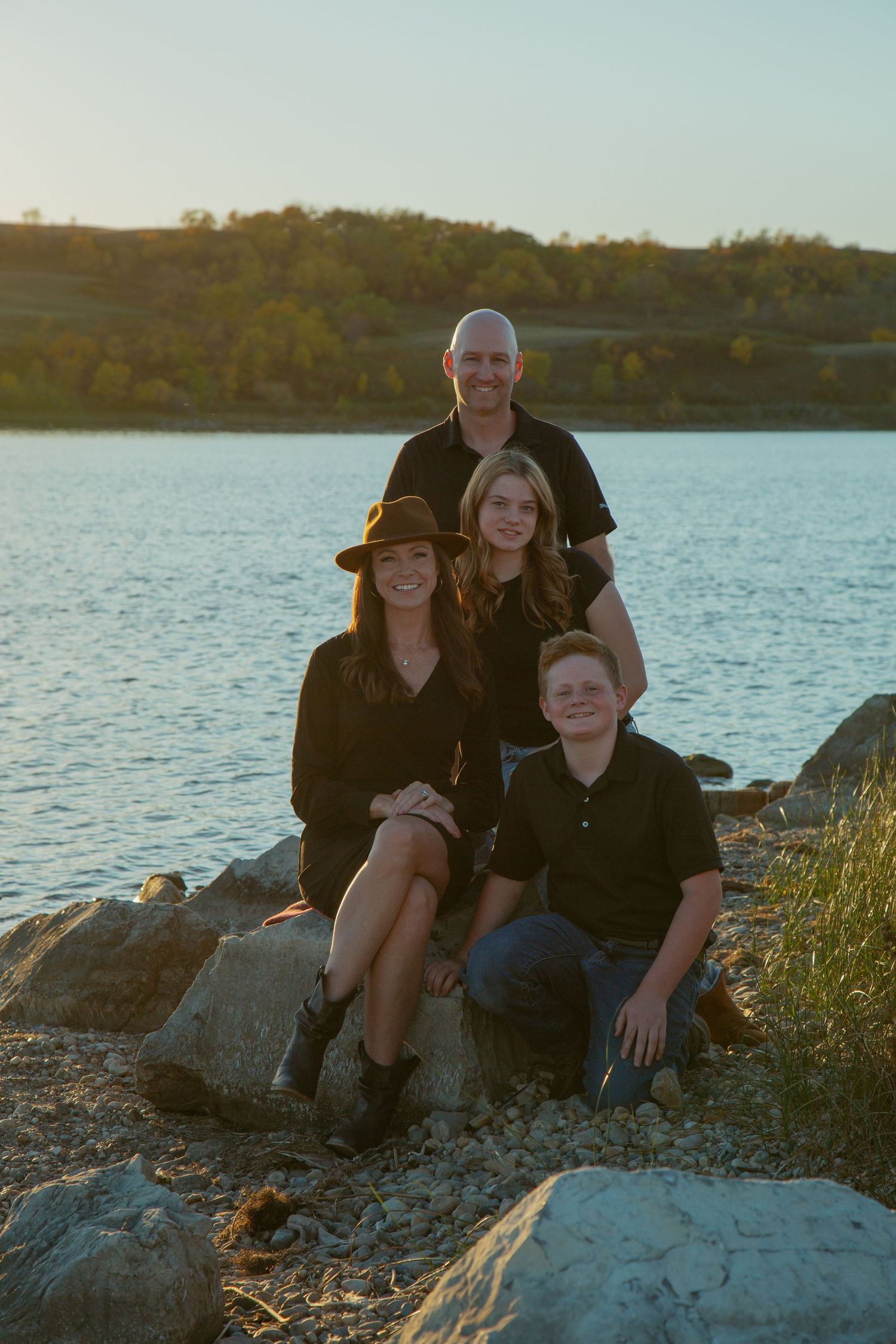 Family group seated on rocky shoreline during sunset at a lake.