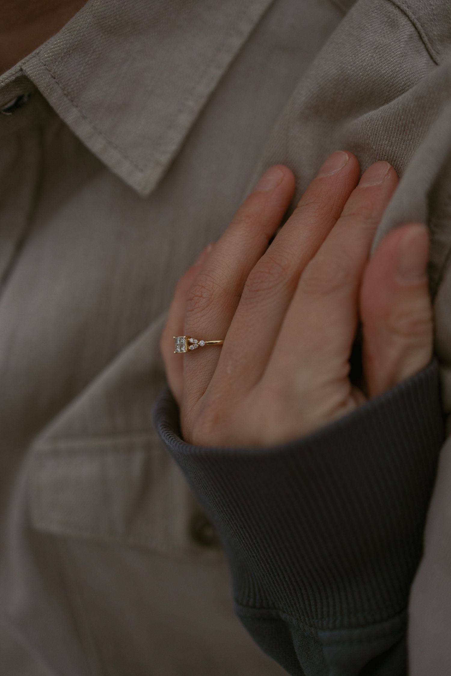 Close-up of a delicate diamond engagement ring on a woman's finger, wearing a beige jacket and grey sleeve.