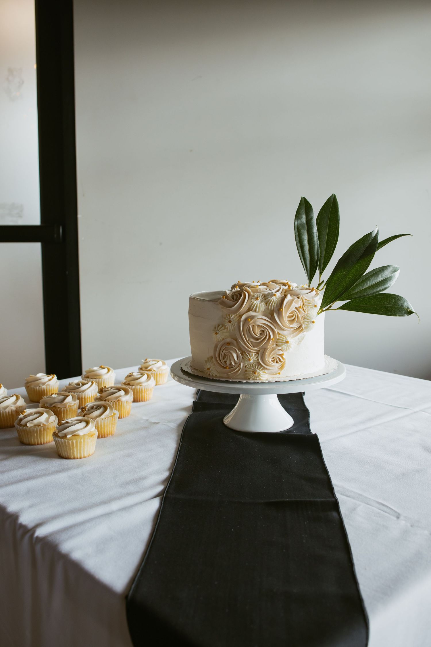 Elegant single-tier wedding cake with cream rosettes and tropical leaf on white stand beside matching cupcakes.