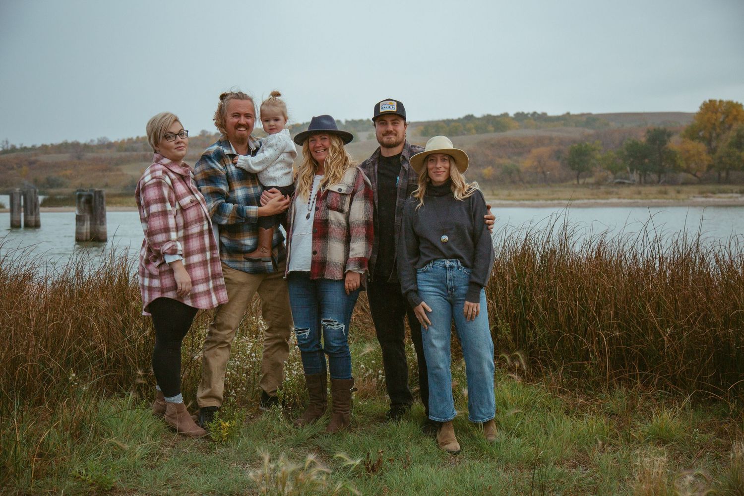 Family group wearing casual fall attire poses together near a lake with autumn foliage in background.