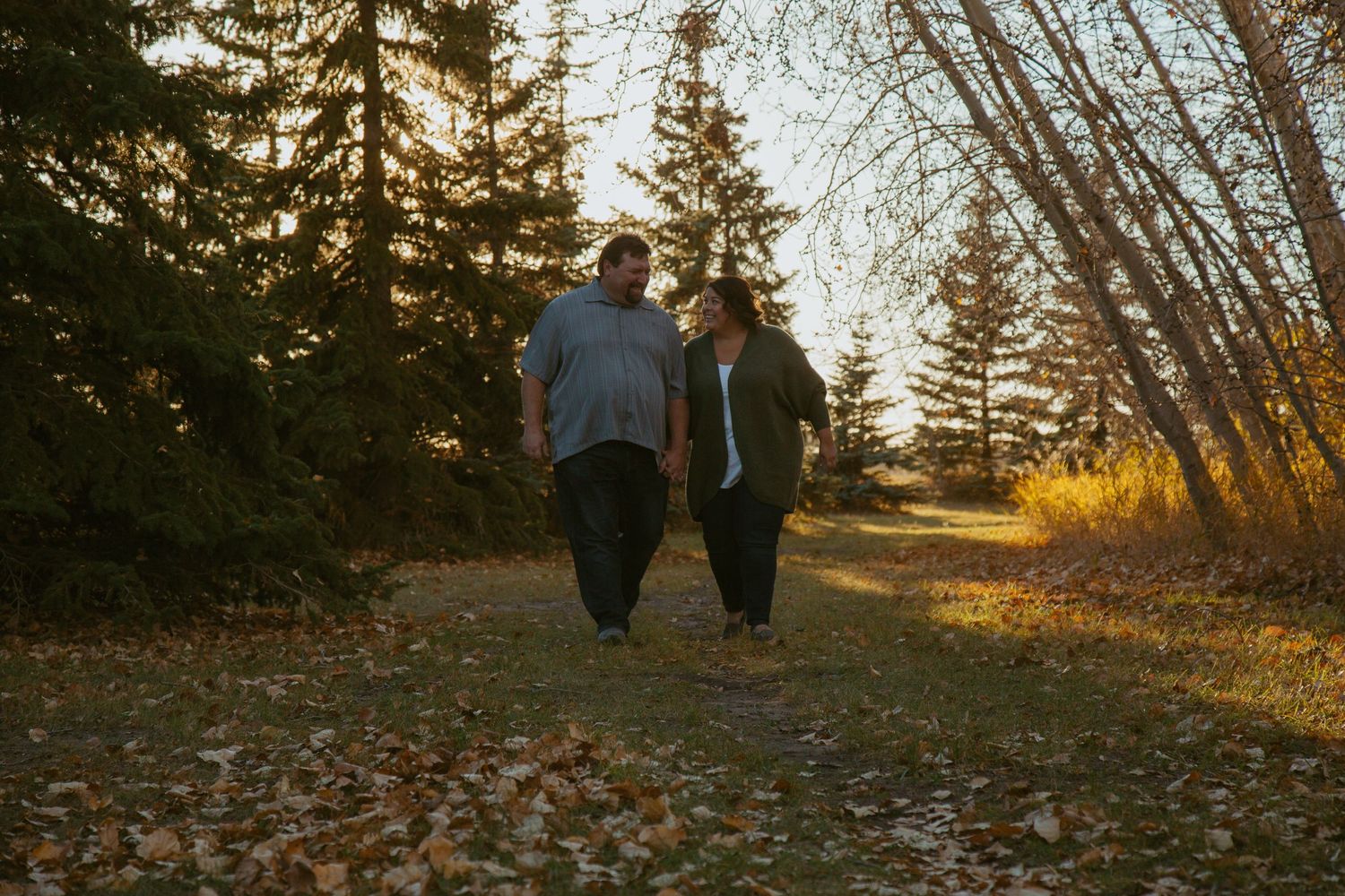 Two people walking together on an autumn path lined with evergreen and deciduous trees.