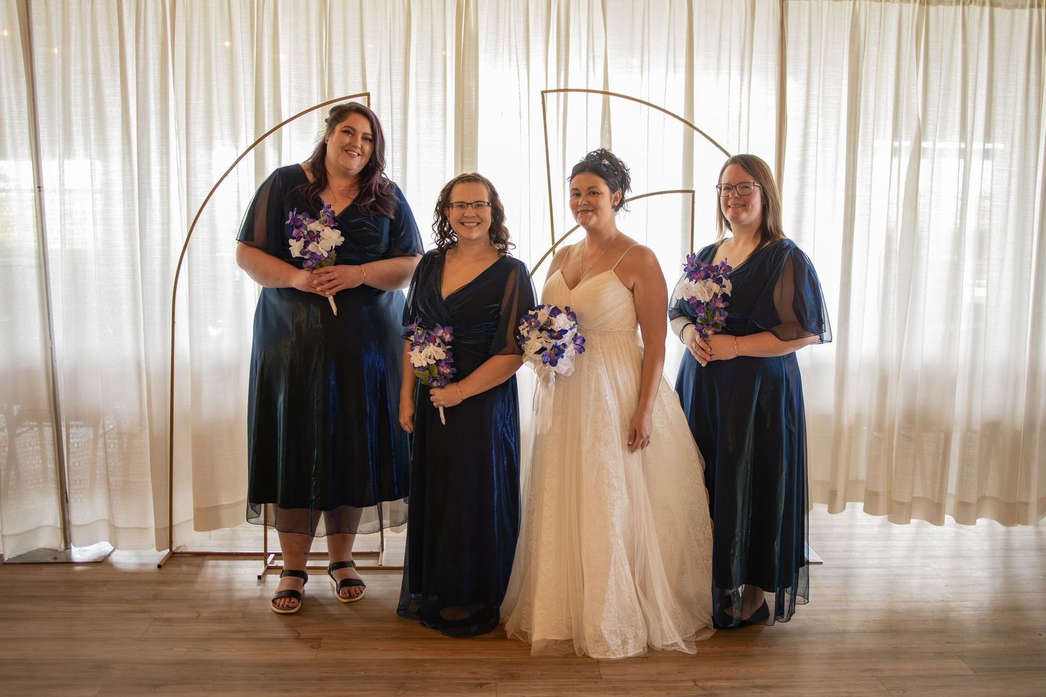 Bride in white gown poses with three bridesmaids in navy blue dresses holding purple bouquets before sheer curtains.