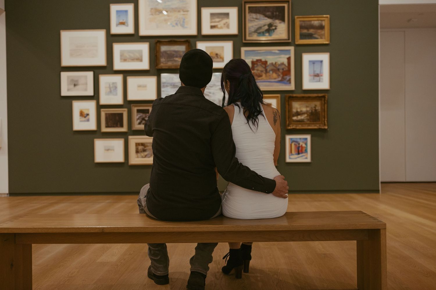 Couple sitting together on a wooden bench in an art gallery, viewing framed artwork on a green wall.