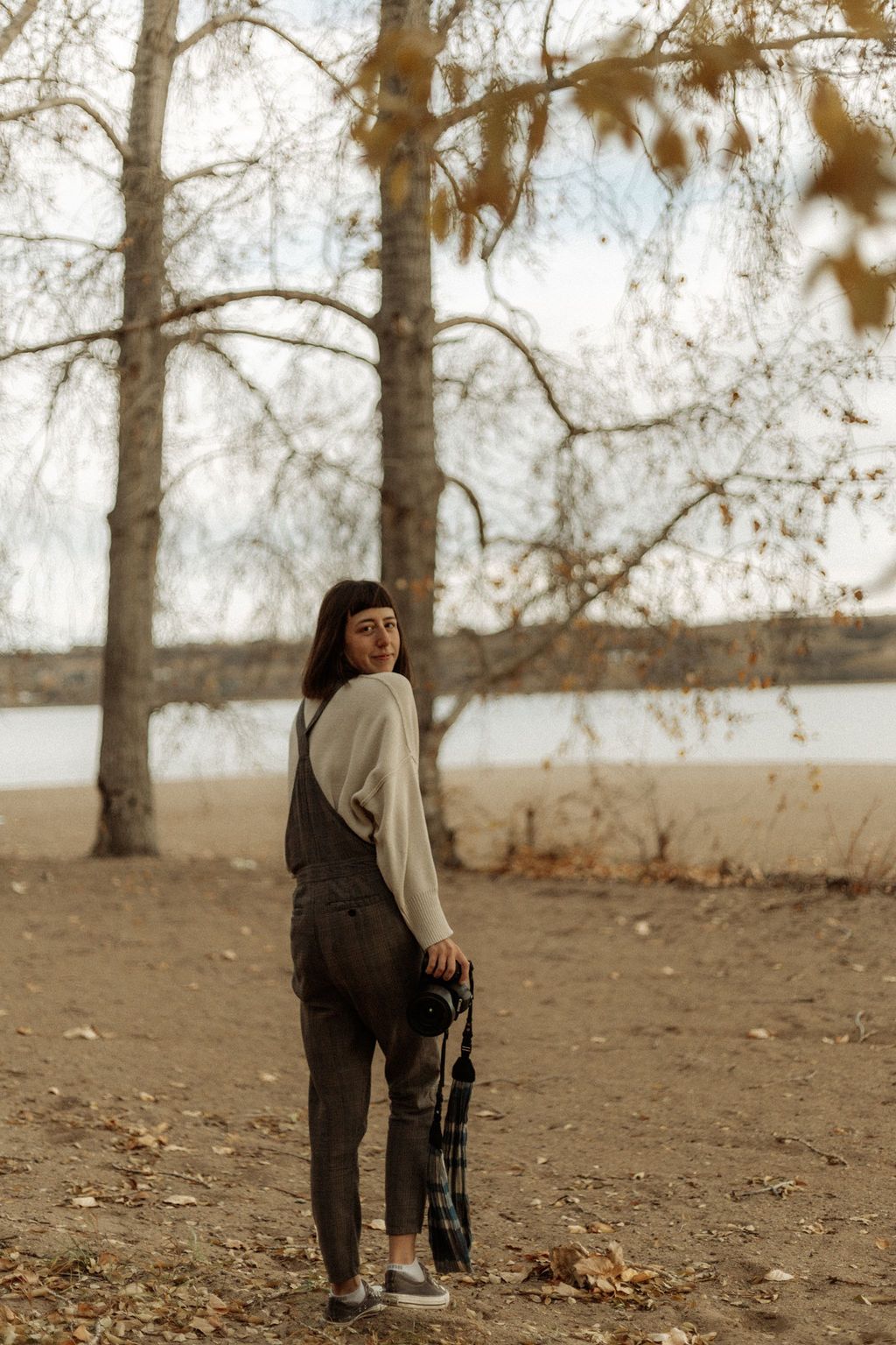 Child wearing overalls explores a wooded area with fallen leaves and bare trees.