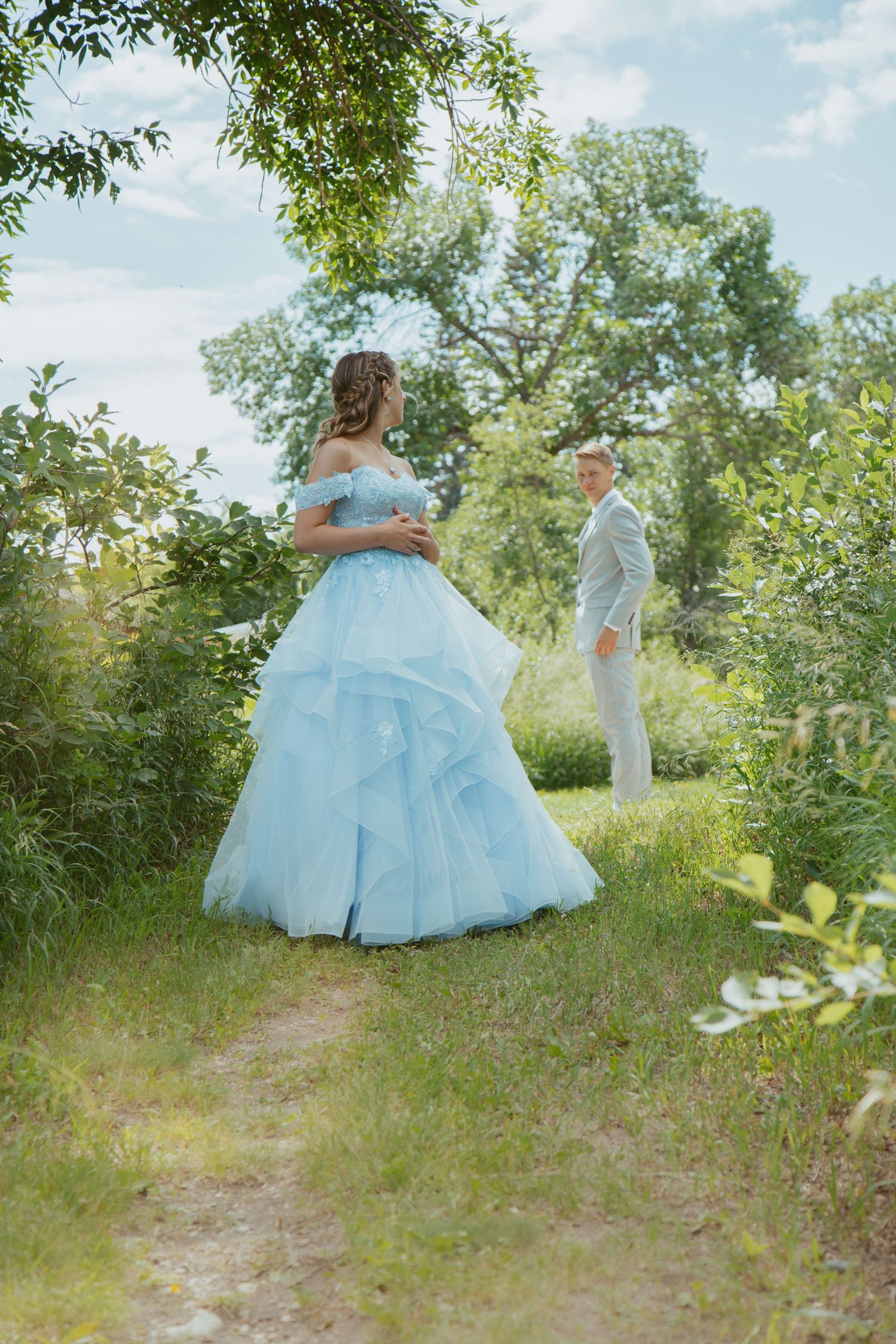 Two formal dresses twirl through a sunlit garden path surrounded by lush green foliage.