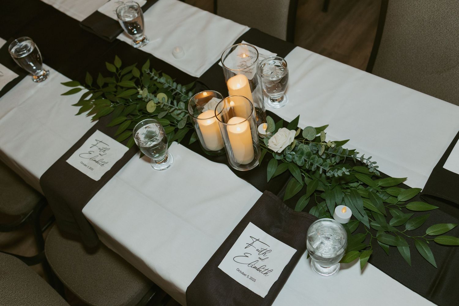 Elegant wedding table with greenery runner, white napkins, calligraphy place cards, and glowing pillar candles.