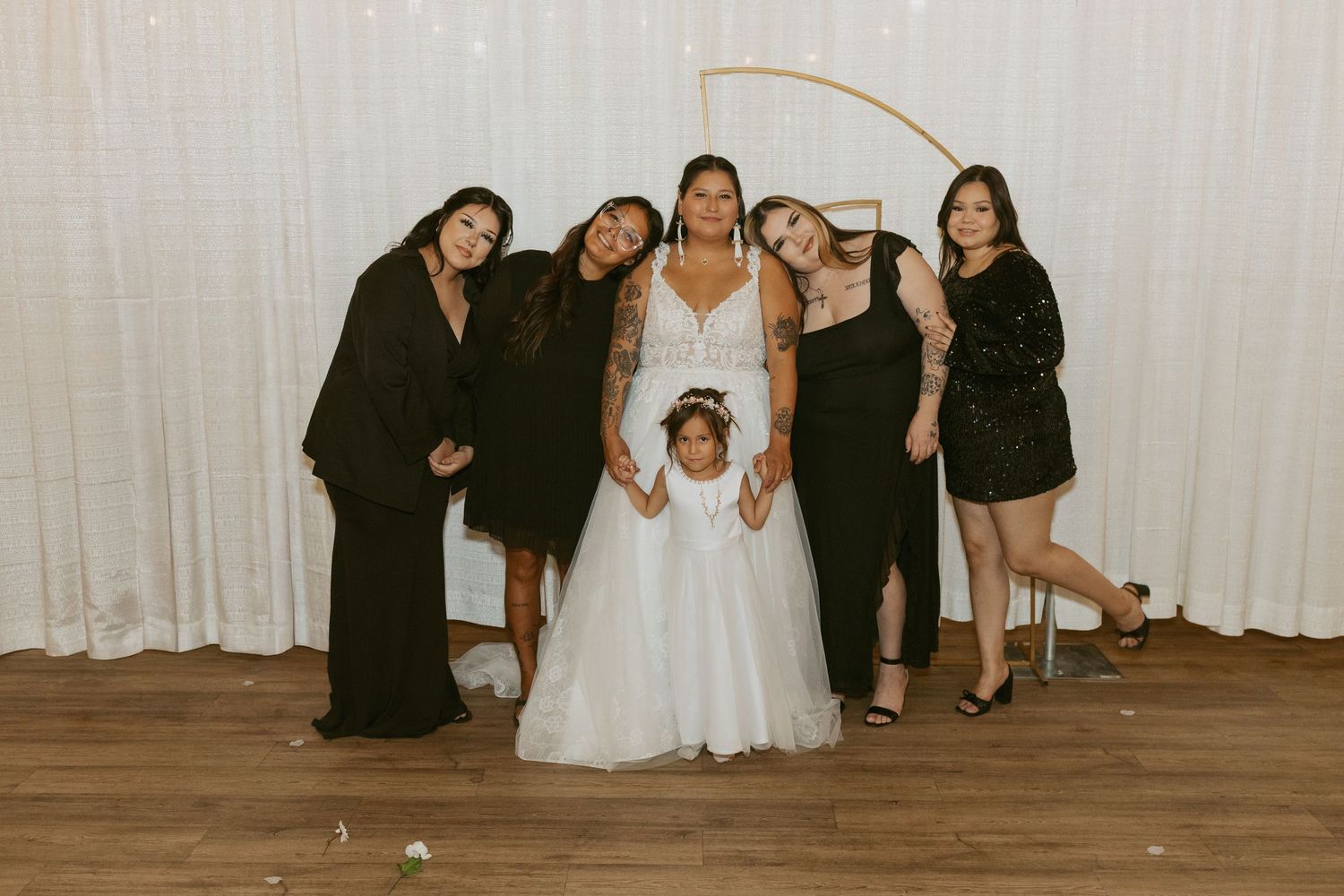 Bride in white gown poses with bridesmaids in black dresses and flower girl at wedding reception.