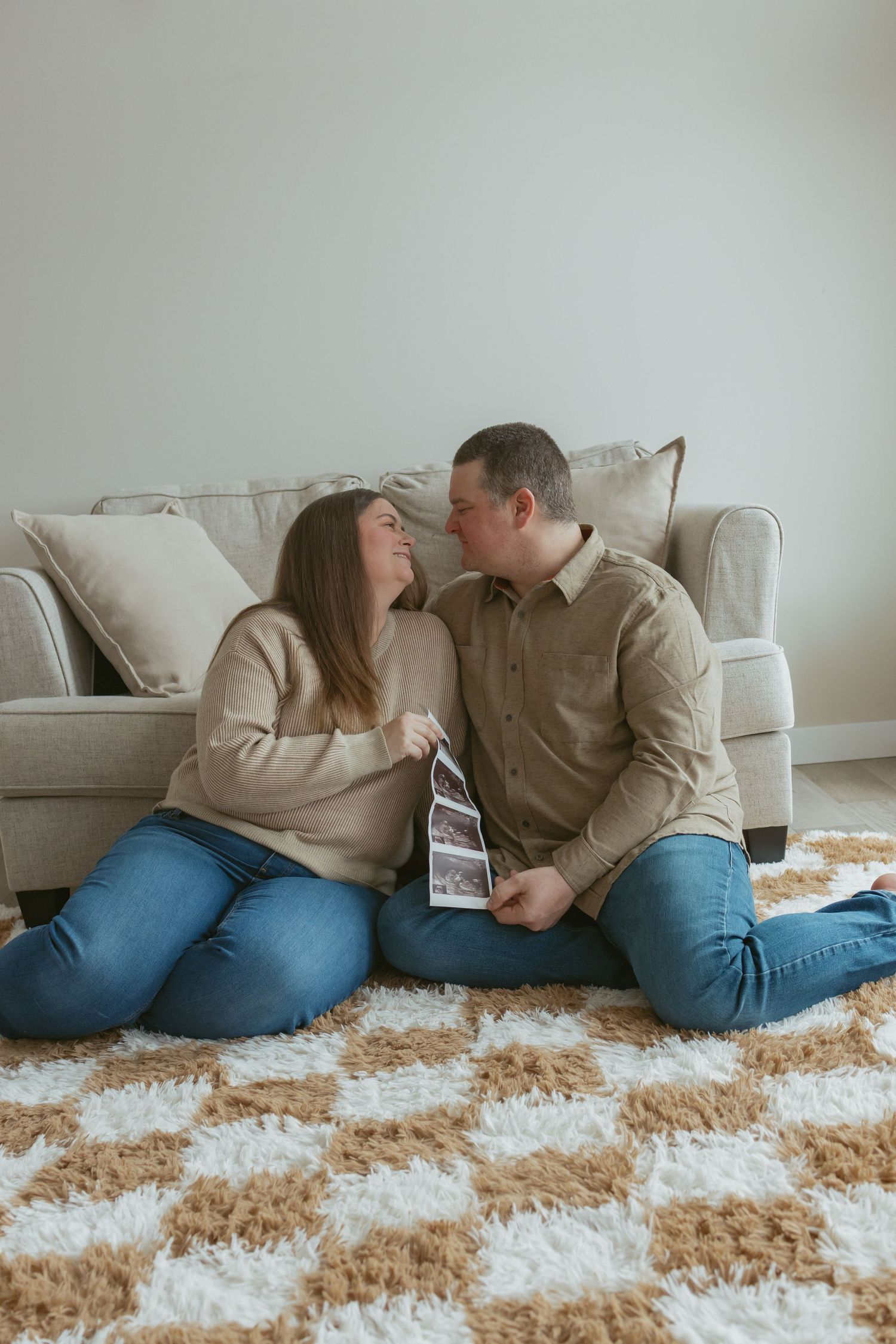 A couple sits on a fluffy rug holding an ultrasound photo, smiling at each other in front of a beige sofa.