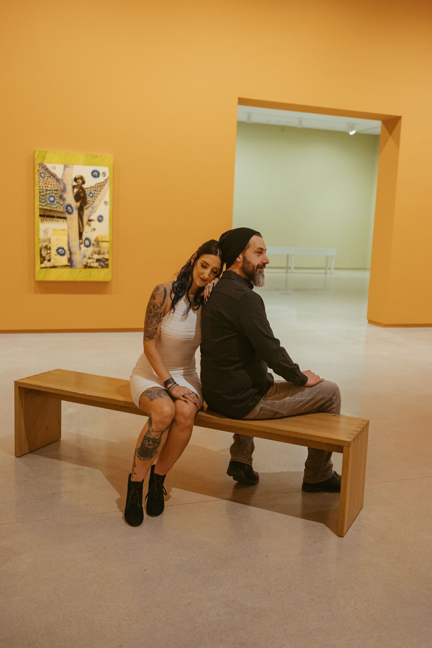 Couple sitting back-to-back on a wooden bench in a yellow art gallery, woman with tattoos smiling.