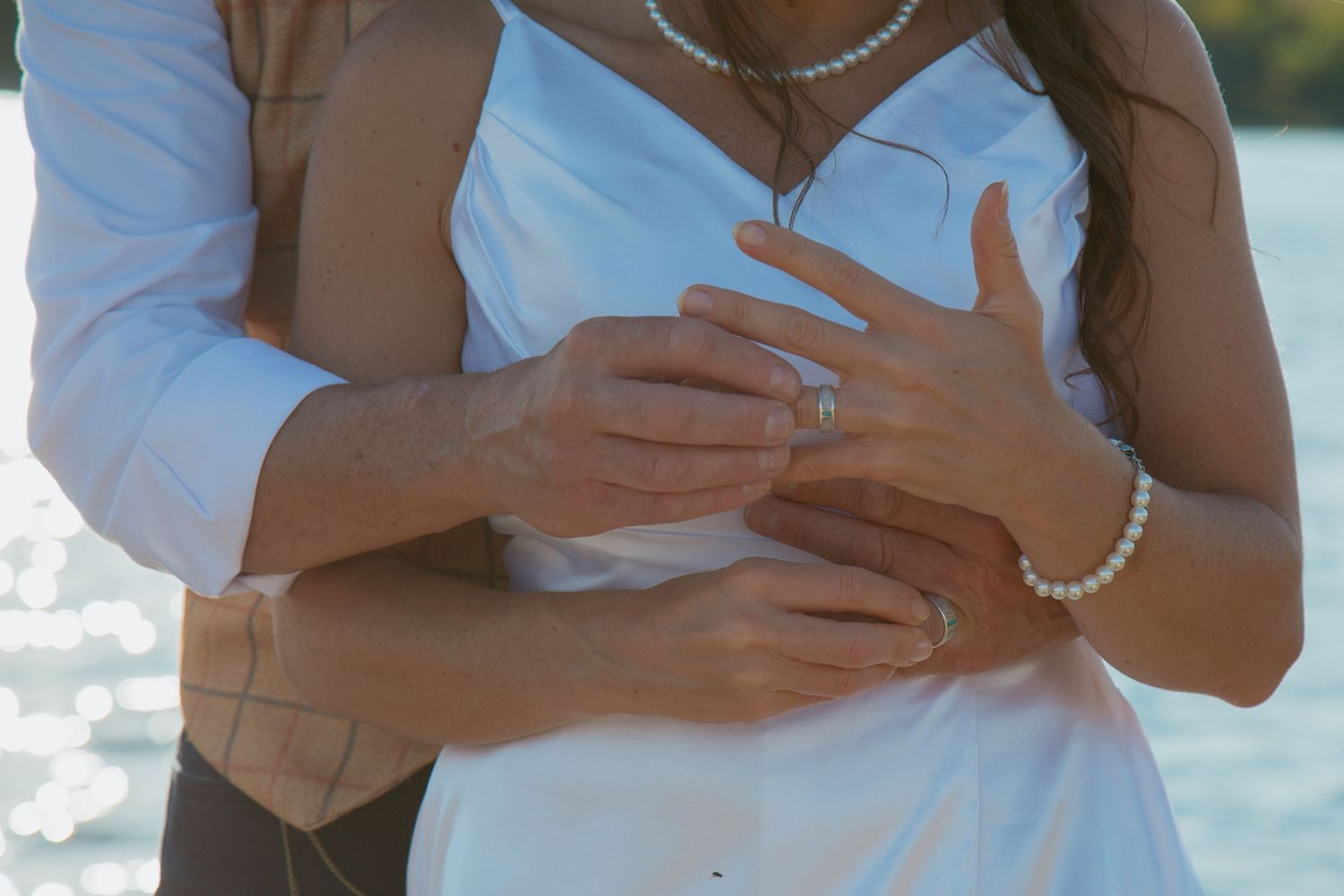 Close-up of hands with wedding rings and pearl bracelet against white dress.