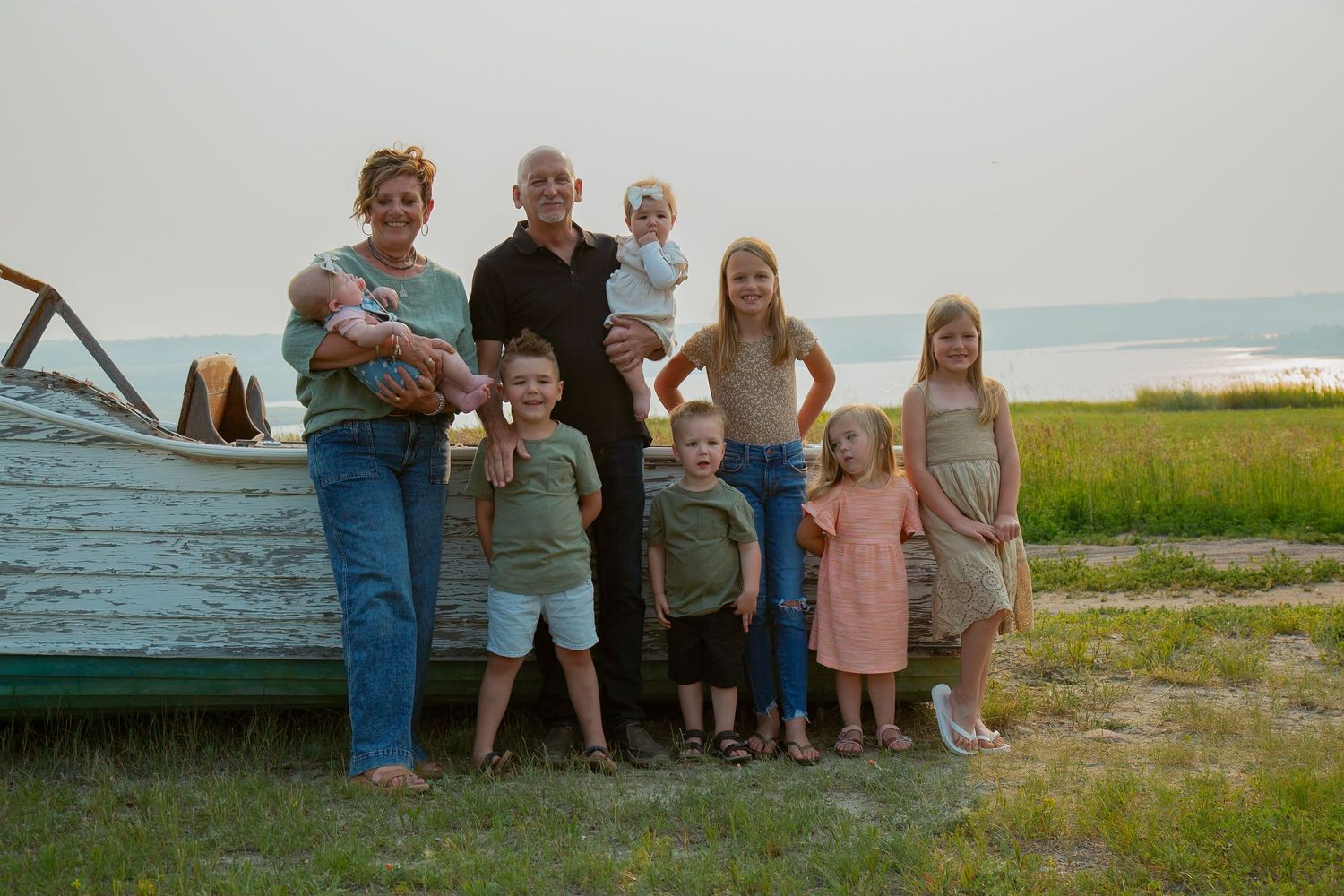 Multi-generational family poses together outdoors near a weathered boat with ocean and grass in background.