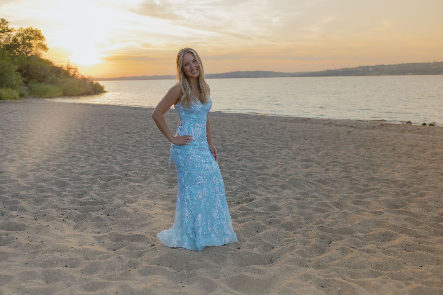 A person in a flowing blue dress walks along a sandy beach during sunset.