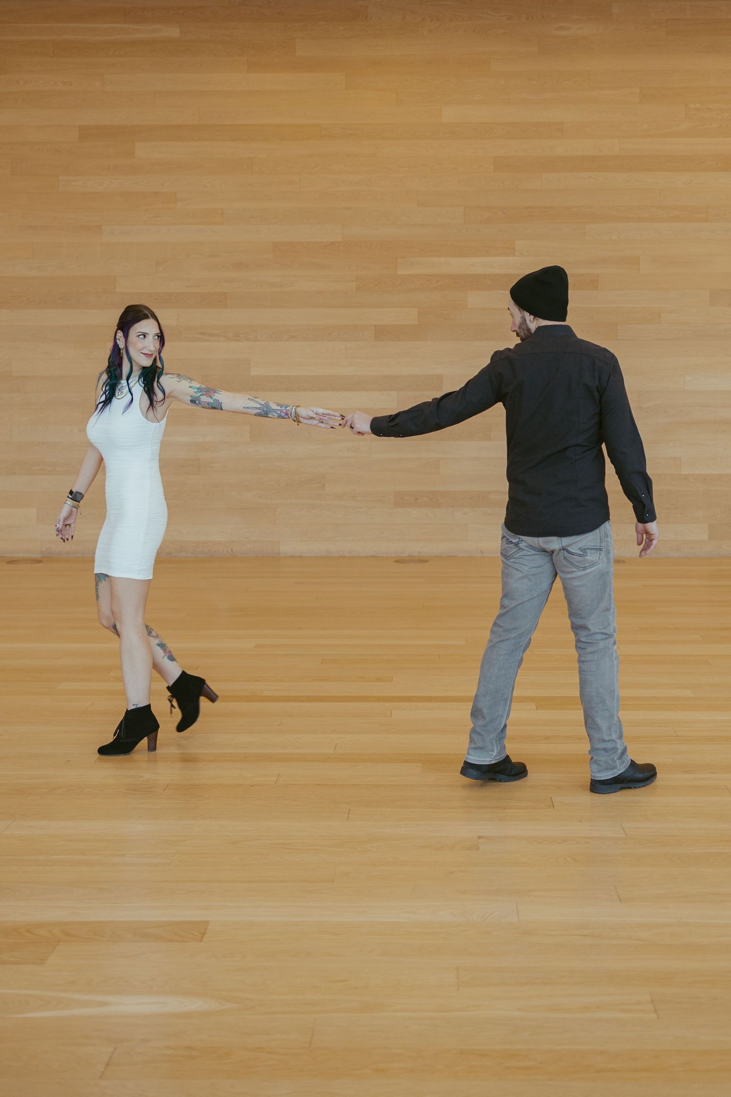 Couple holding hands on a hardwood dance floor, woman in white dress with tattoos and black boots.
