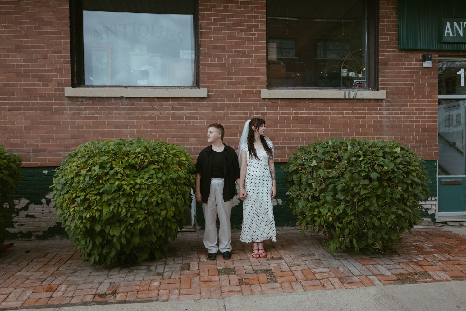 A couple stands together outside a brick building with greenery.