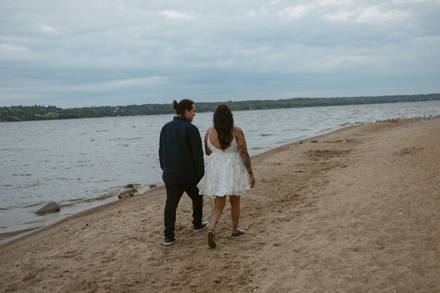 A couple walks along a sandy beach shoreline on an overcast day.