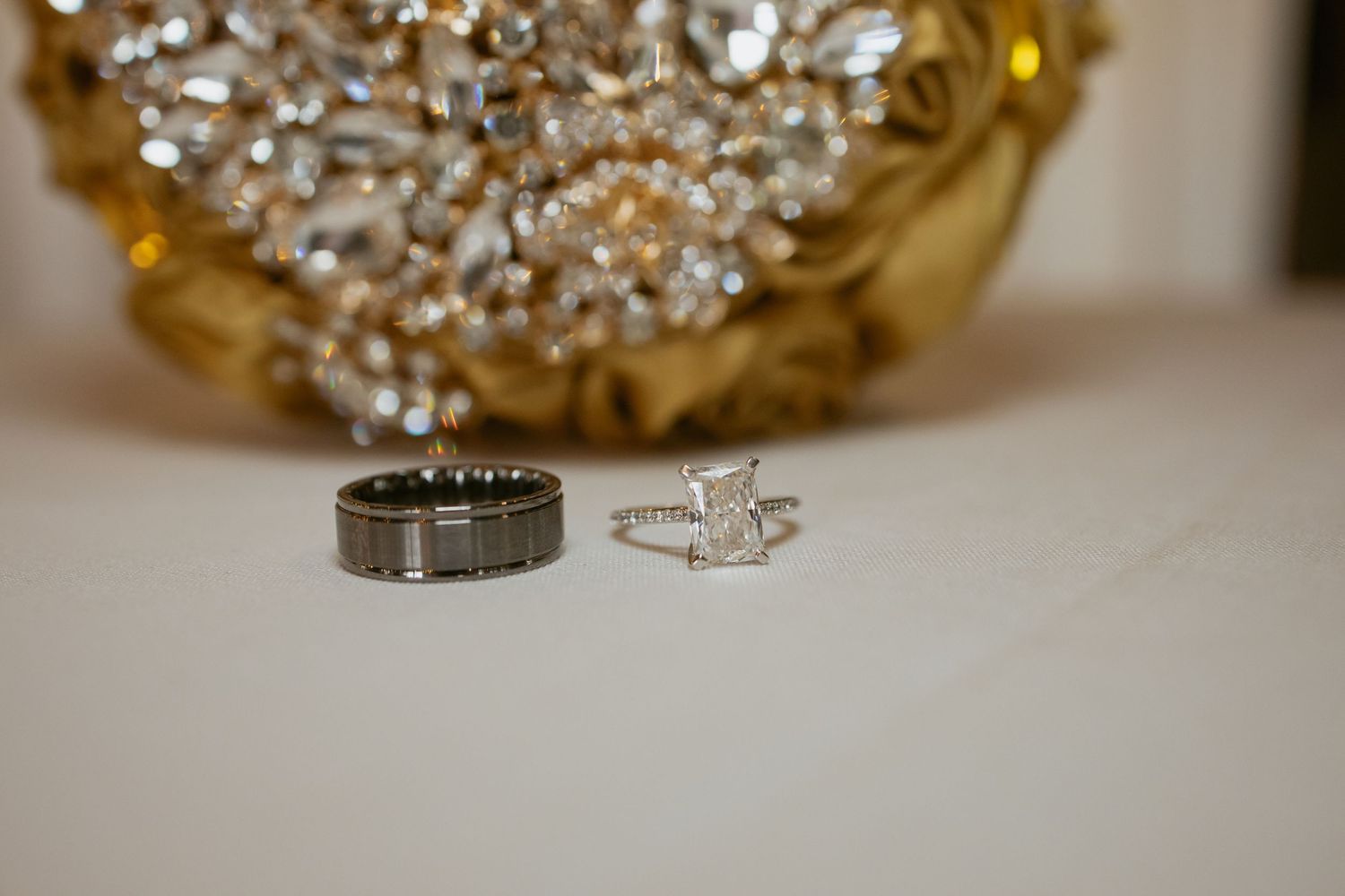 Silver wedding band and diamond engagement ring displayed on white surface with gold chandelier bokeh background.