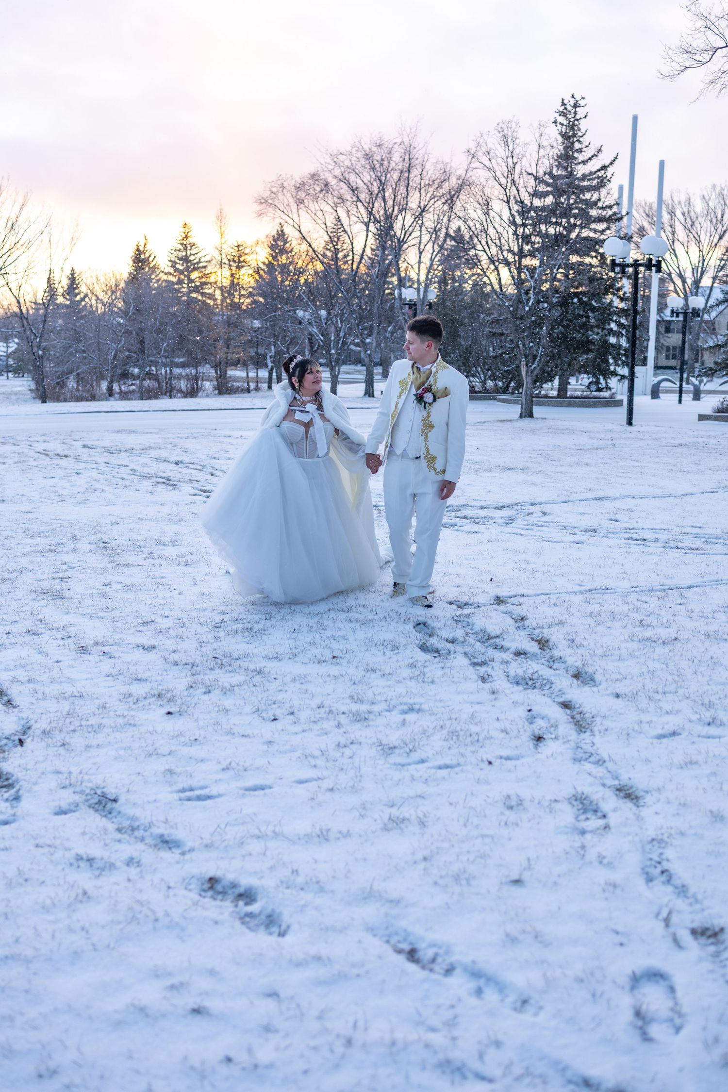Bride and groom holding hands walking through snowy winter park at sunset in white wedding attire.