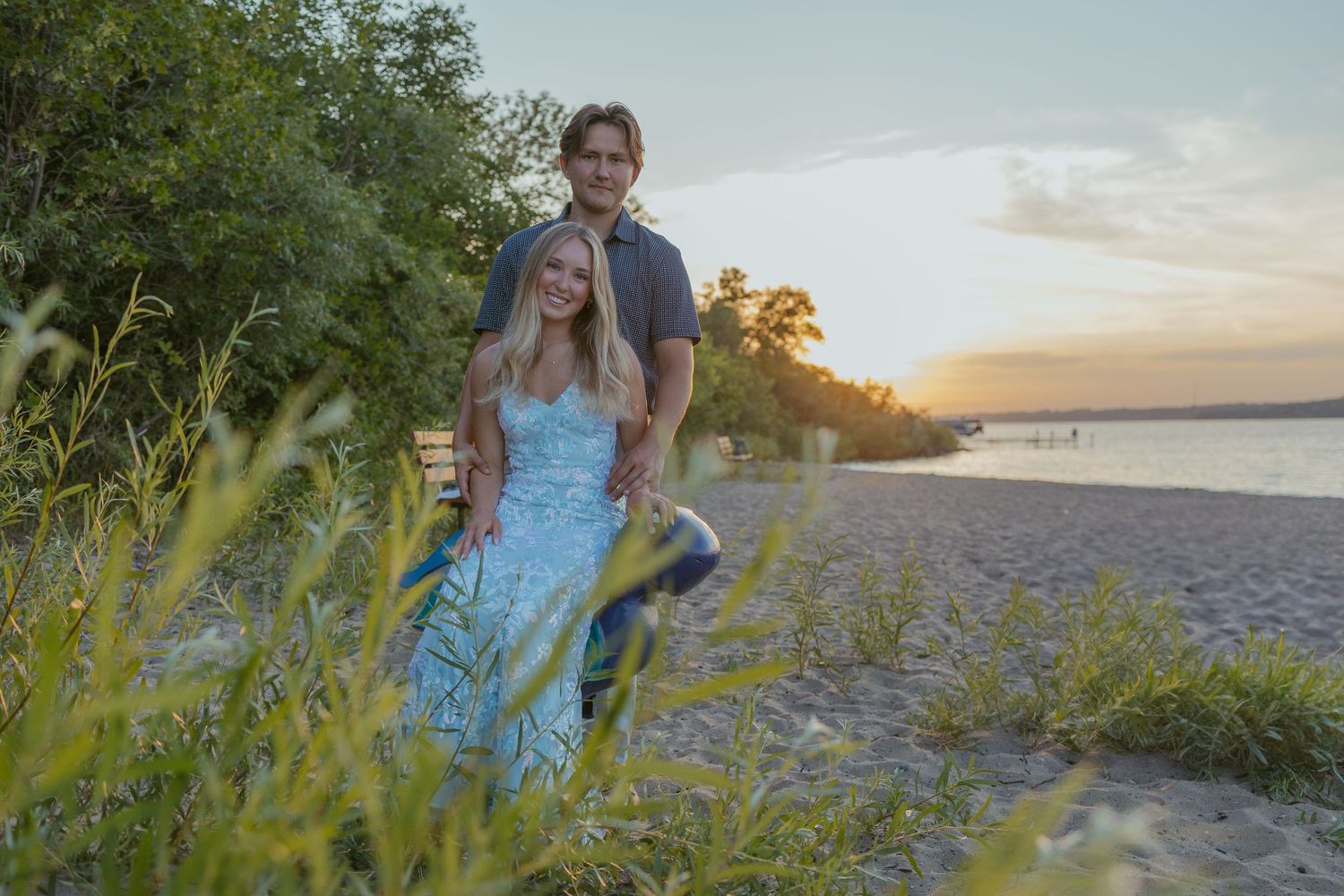 Two people walk together on a beach path surrounded by greenery at sunset.