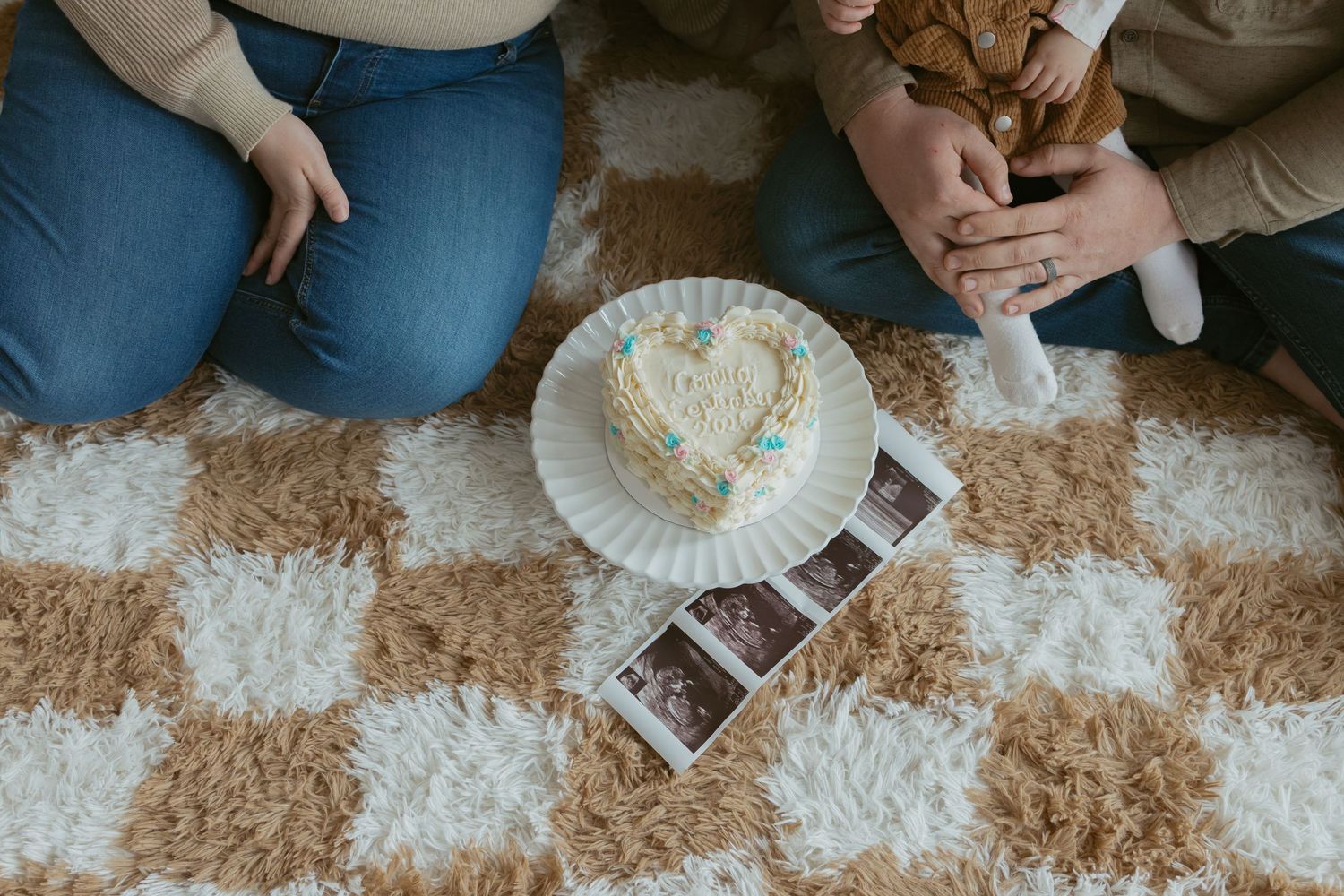 Couple sitting on checkered rug with heart-shaped cake and ultrasound photos for a pregnancy announcement.