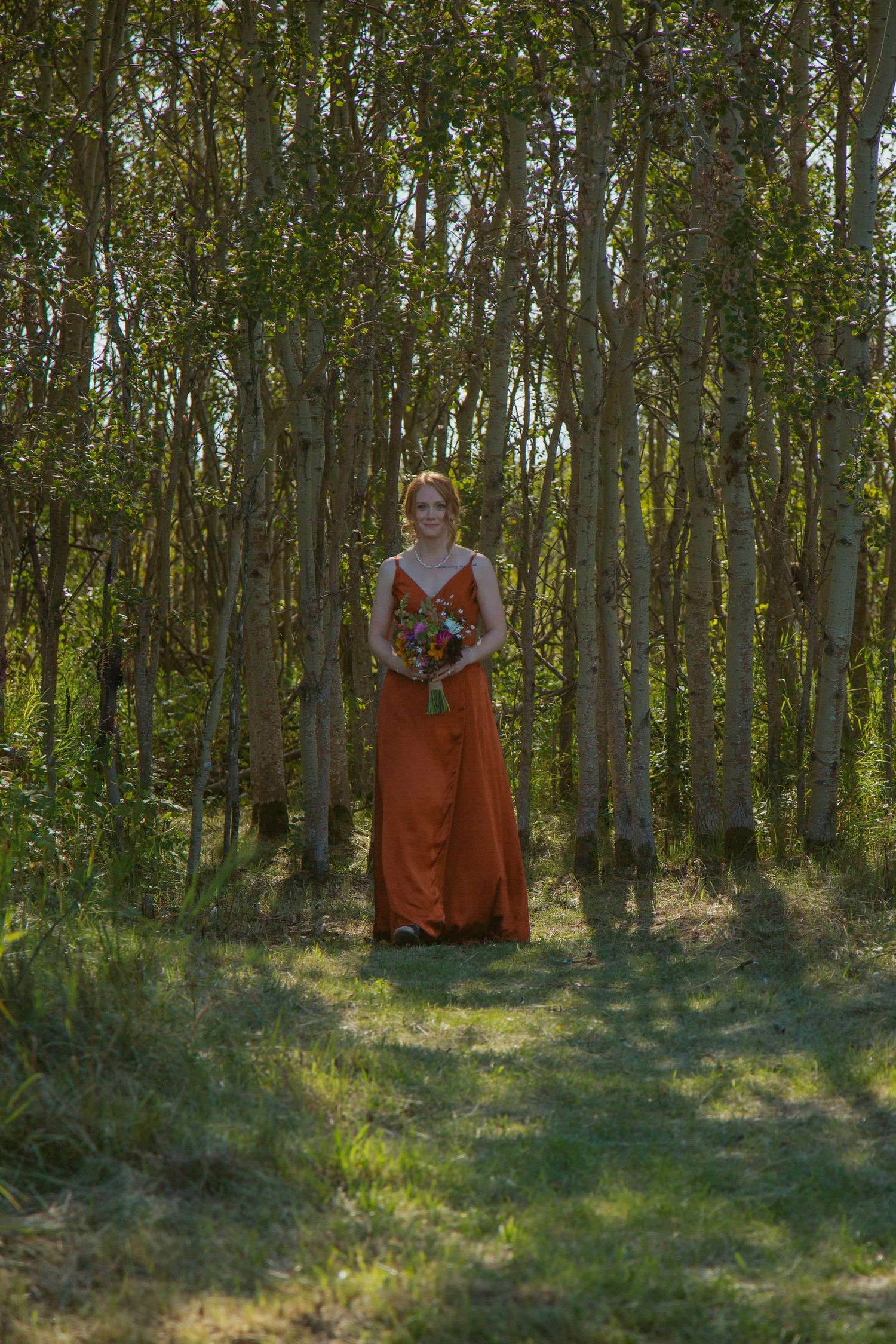 Person in orange dress poses in a serene forest setting with filtered sunlight.