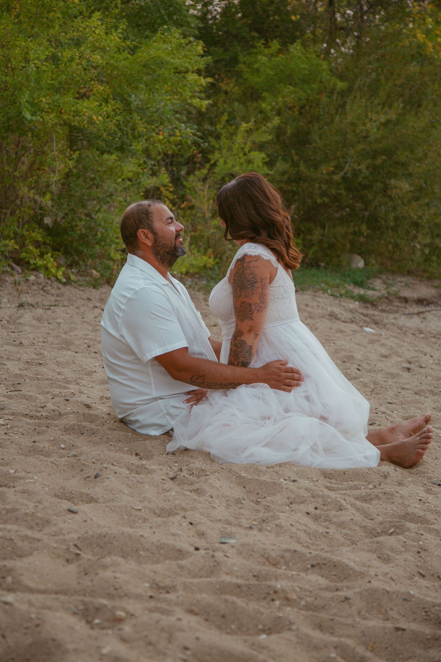 Couple sitting together on a sandy beach with greenery in the background.