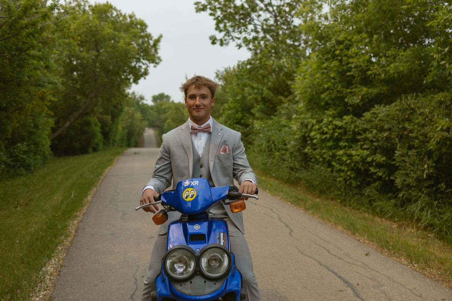 Person rides blue motor scooter on tree-lined path during summer.