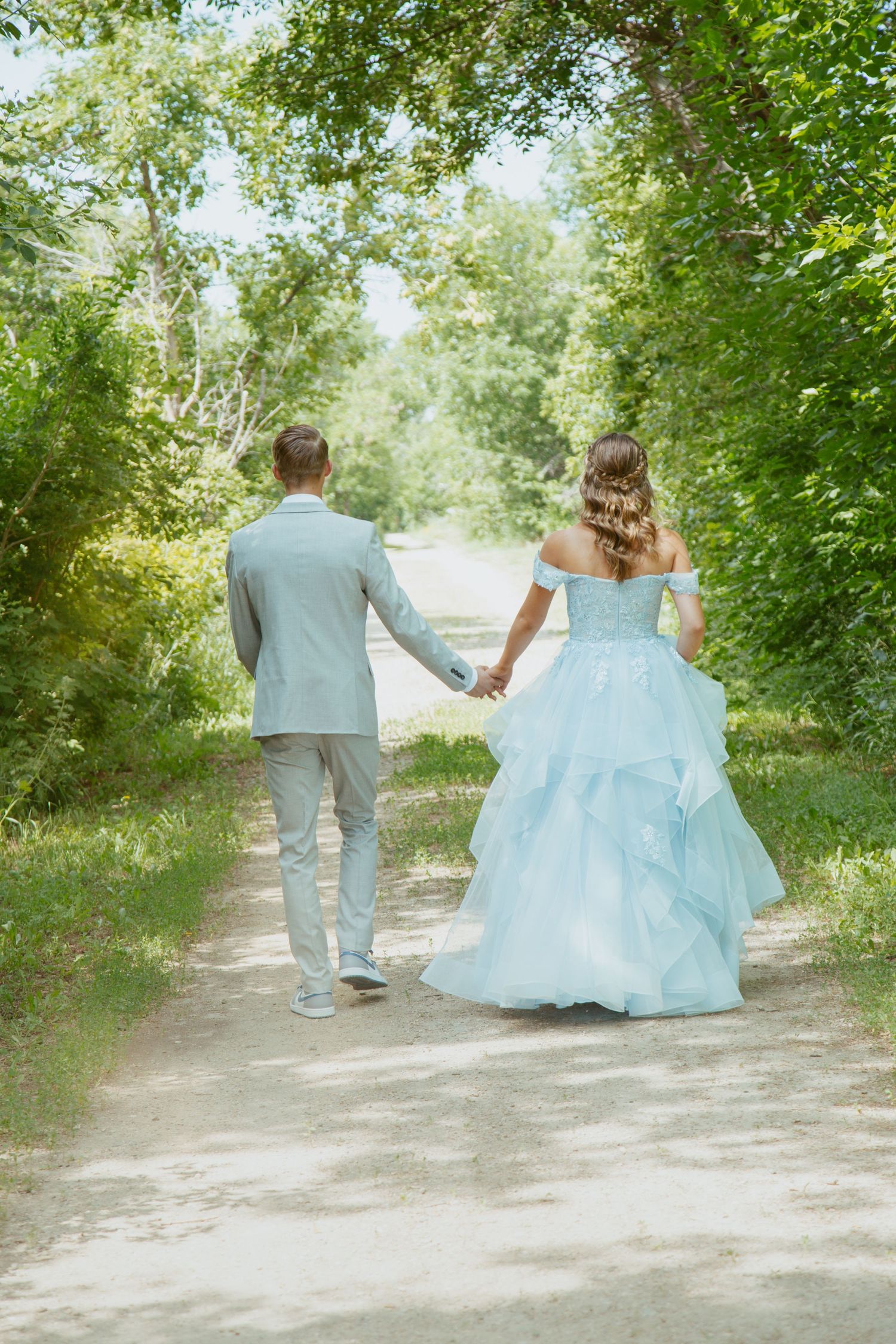 Two people walk down a tree-lined path, one wearing a grey suit and another in a light blue ballgown.